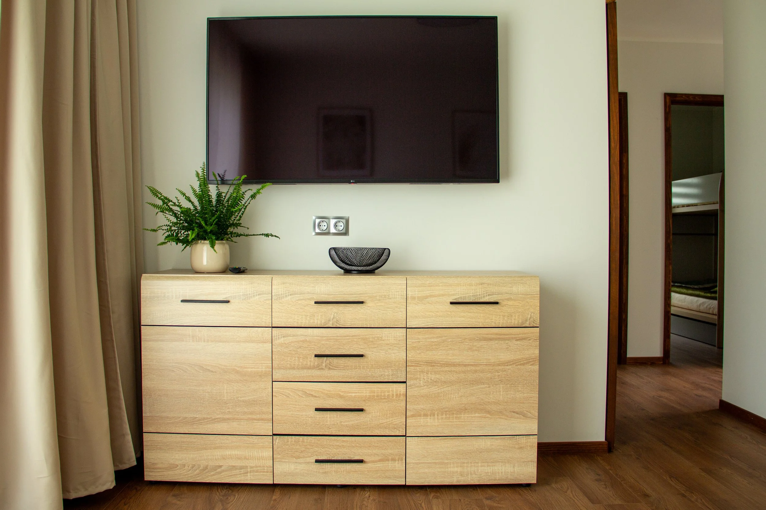 Wooden dresser with black handles, decorated with a potted plant, a black bowl, and a black remote, against a white wall with a large flat-screen TV mounted above, in a room with cream curtains and wooden flooring.