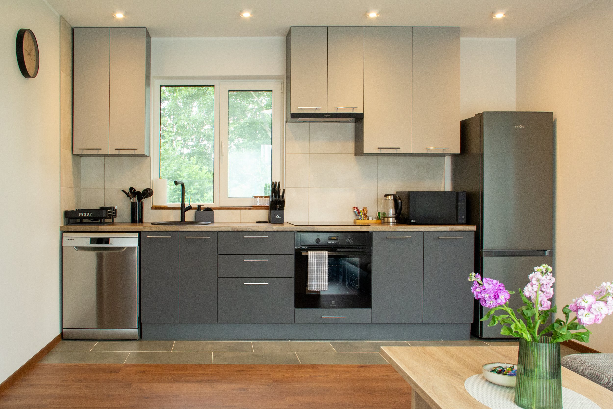 Modern kitchen with grey and white cabinets, a black oven, refrigerator, microwave, and kettle. There is a window above the sink with green trees outside. A vase with pink and purple flowers is on a wooden table in the foreground.