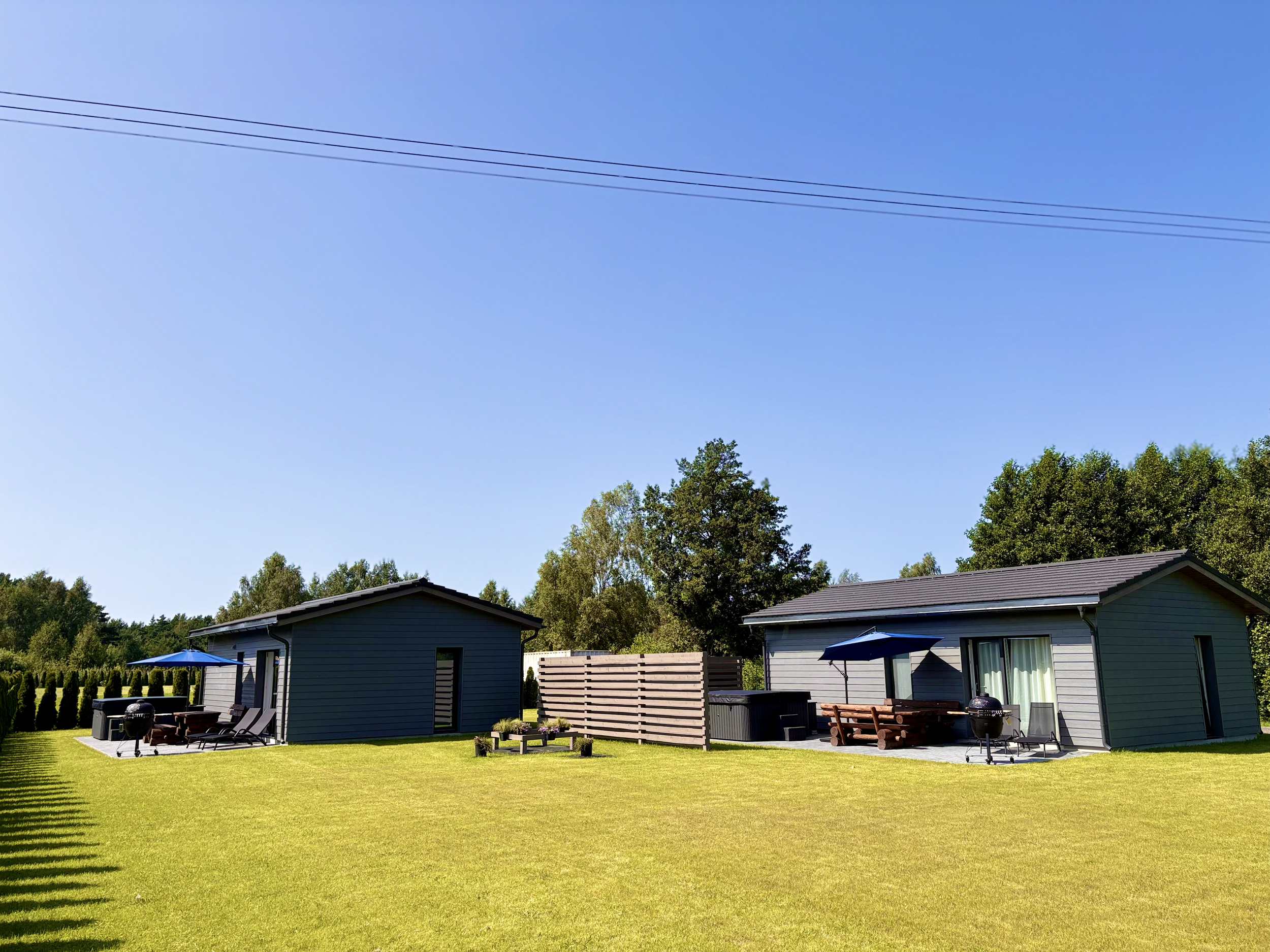 Two gray cottages with outdoor patios, umbrellas, and grills in a grassy yard, surrounded by trees under a clear blue sky.