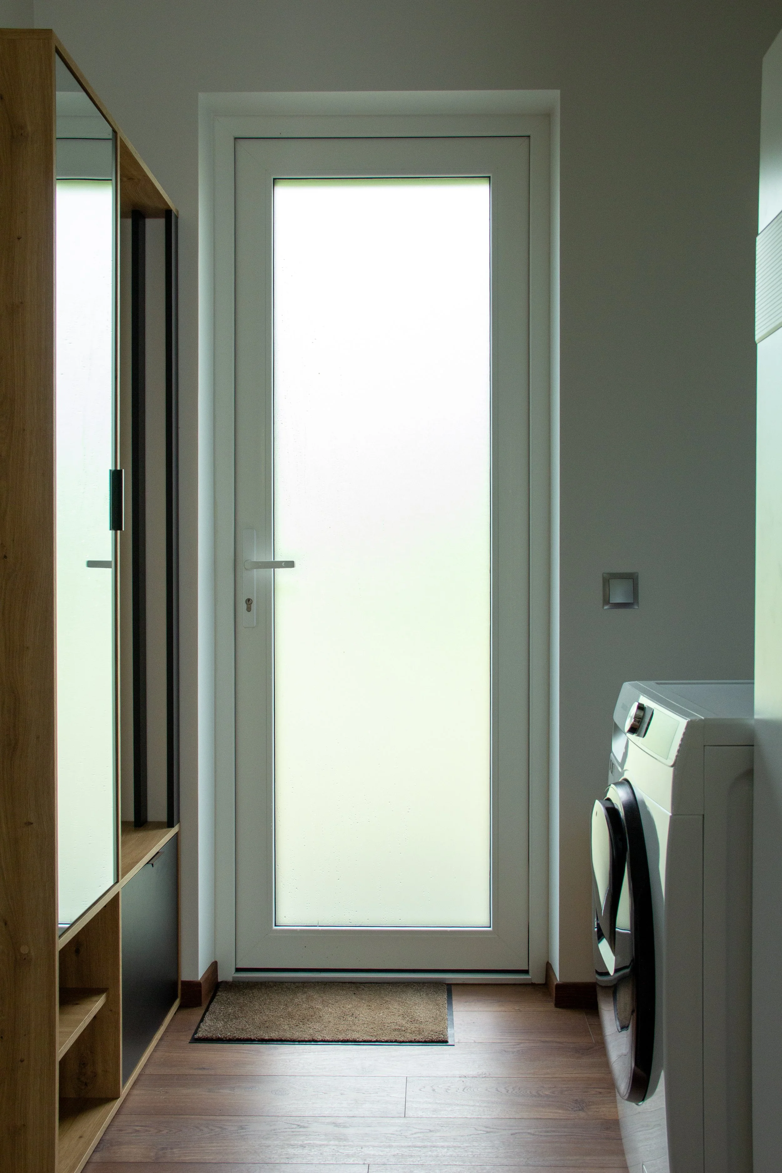 Interior view of a narrow laundry room with a frosted glass door, wooden floor, a black doormat, a wooden shelving unit with mirrors, and a white washing machine.