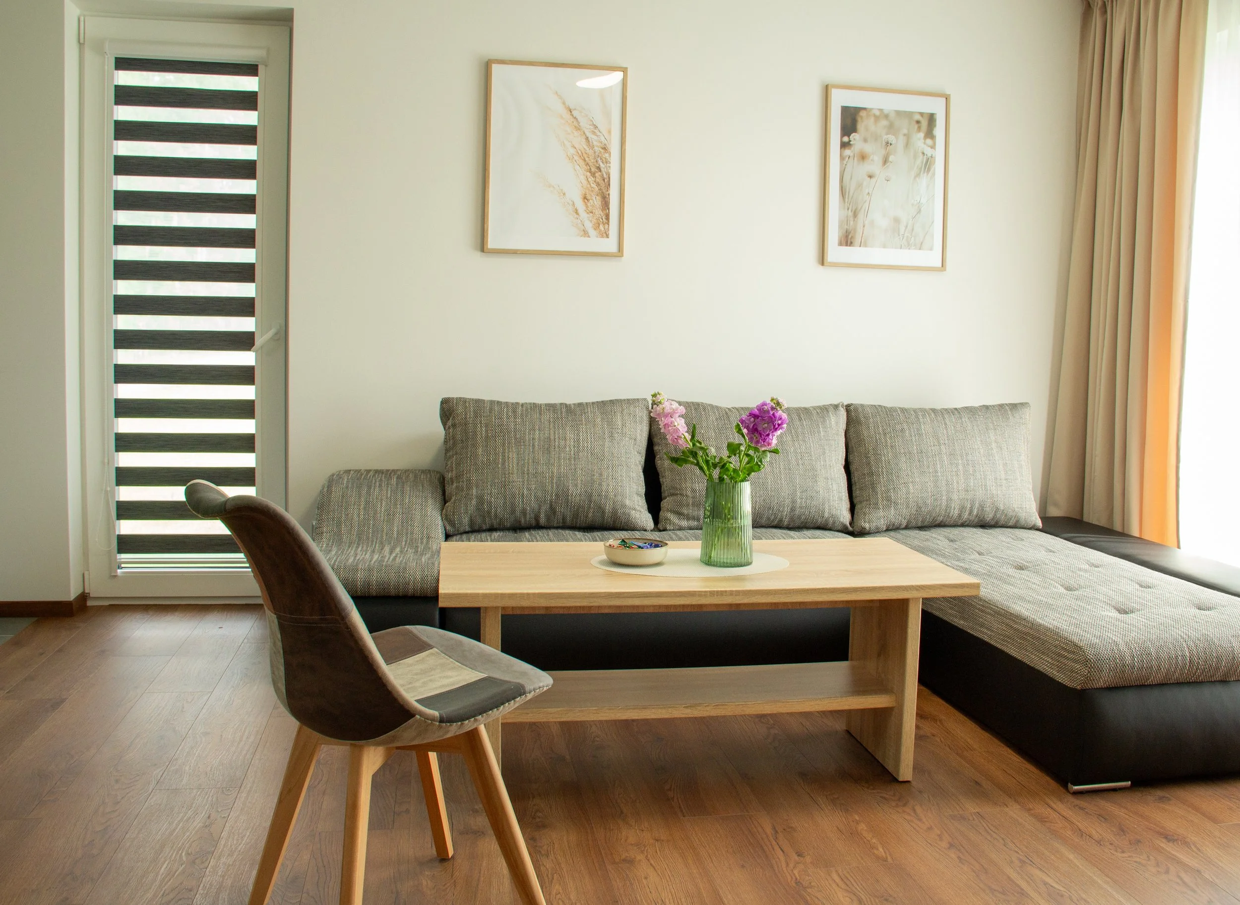 Living room with a gray sofa, a wooden coffee table with a vase of purple flowers, framed artwork on the wall, and a chair in front of the table.