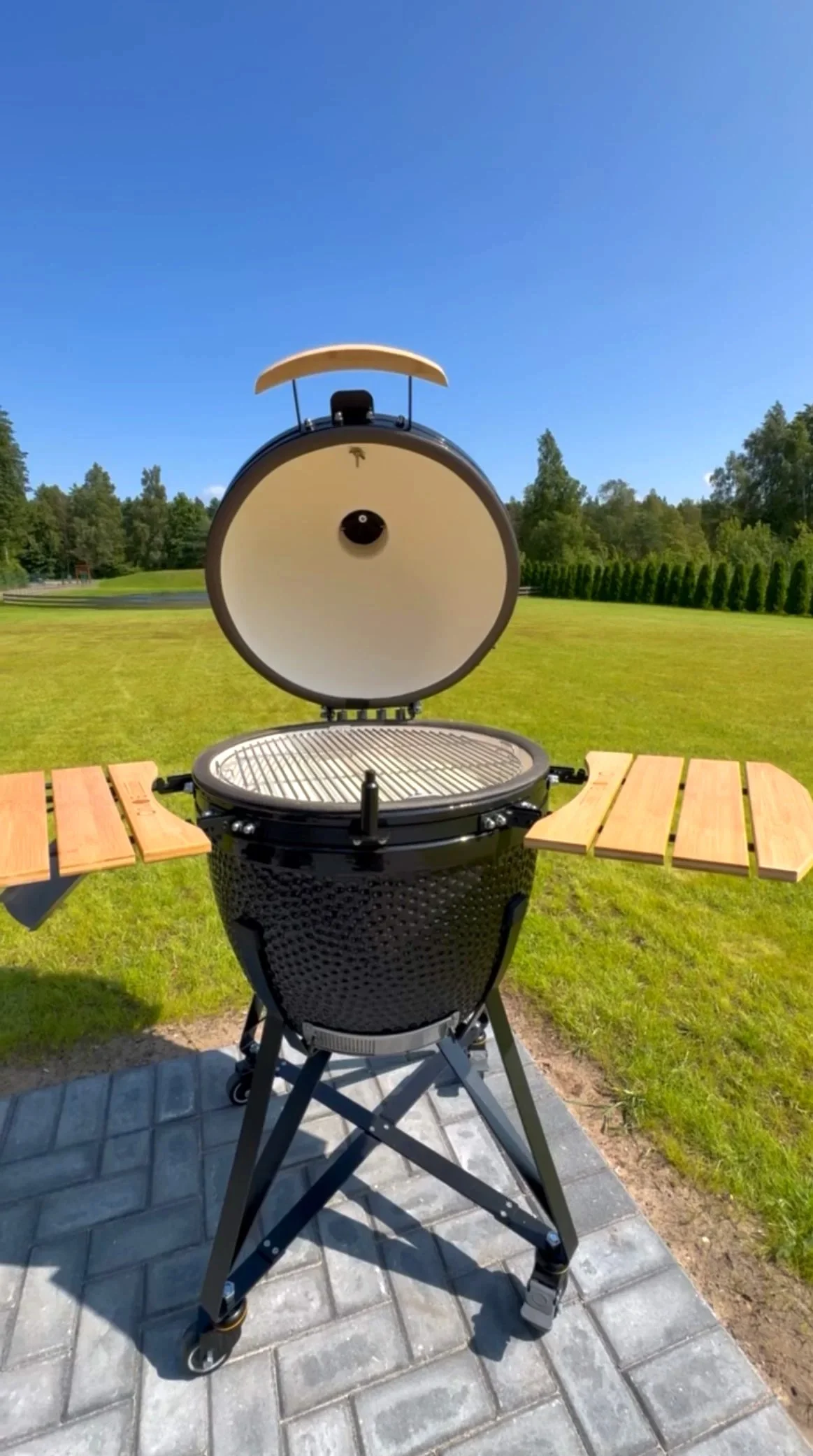 A black grill with a lid open, revealing the cooking grate inside, sitting outdoors on a paved surface with grassy fields and trees in the background under a blue sky.