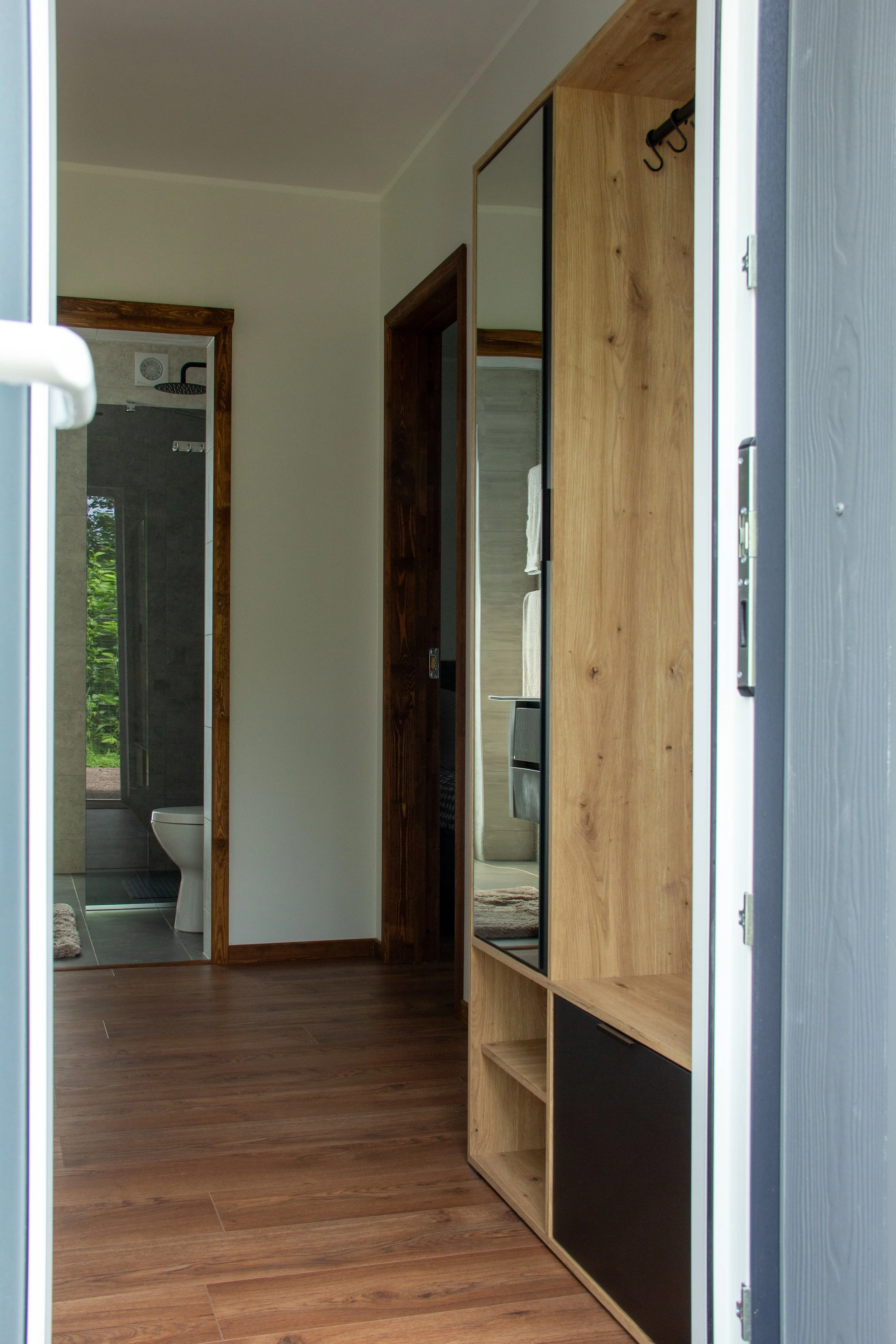 Interior view of a modern home hallway showing a wooden storage unit with a mirror, with an open door leading to a bathroom with a visible toilet and a walk-in shower.