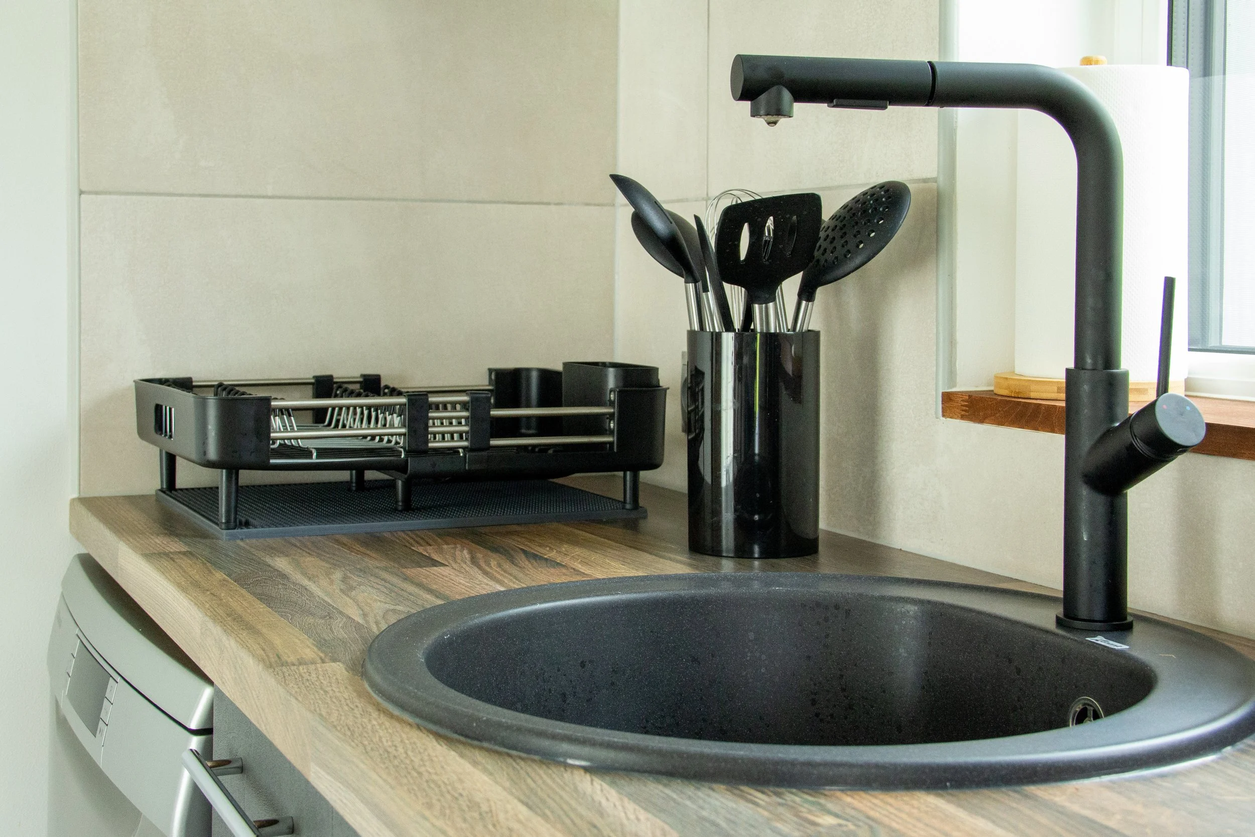 Kitchen countertop with black sink, black kitchen utensils in a black container, dish rack, and a window in the background.