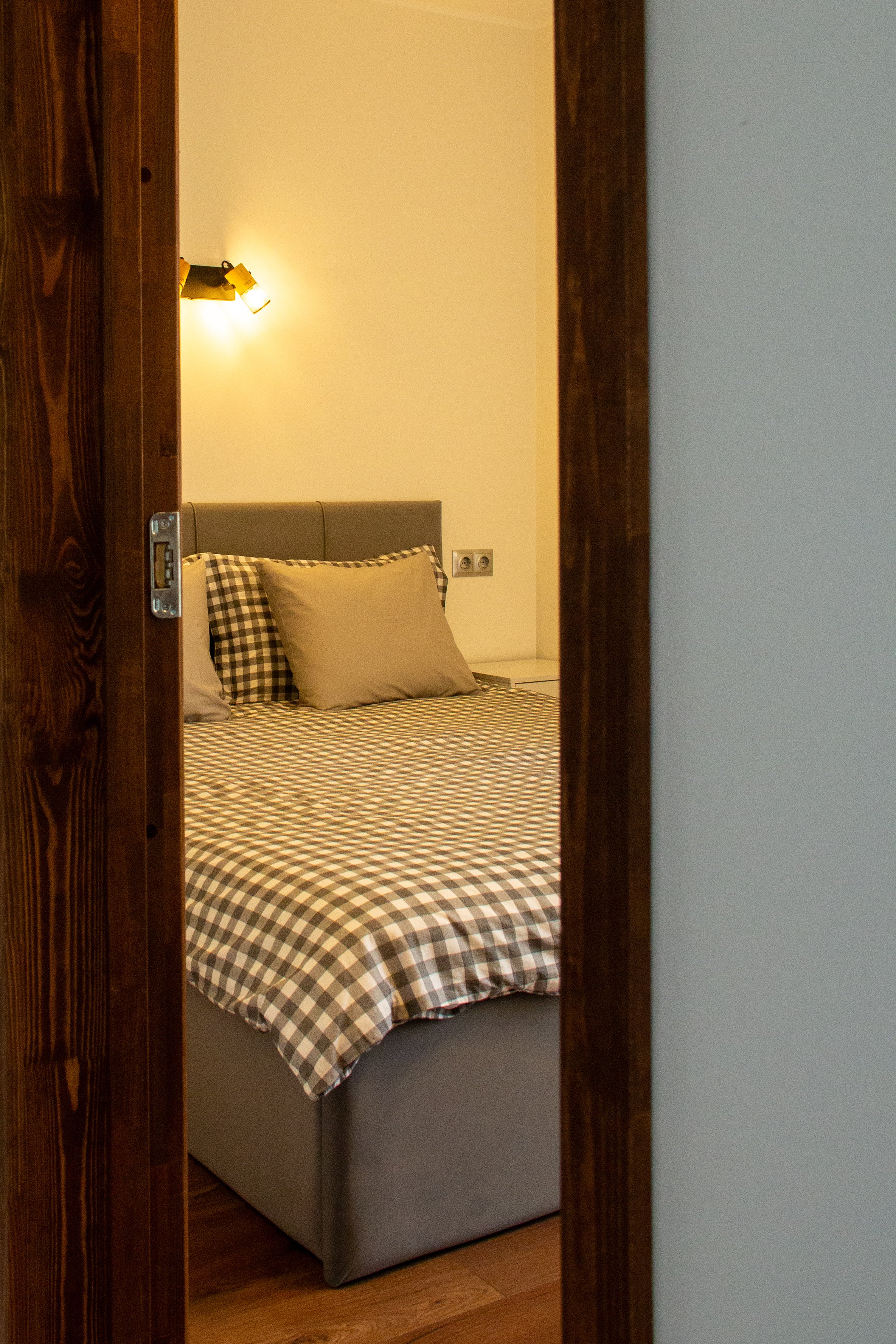 A bedroom with a neatly made bed. The bed has a checkered bedspread and three pillows. The headboard is upholstered, and there's a wall-mounted light fixture above. The door frame is wooden, and the room's walls are painted white.