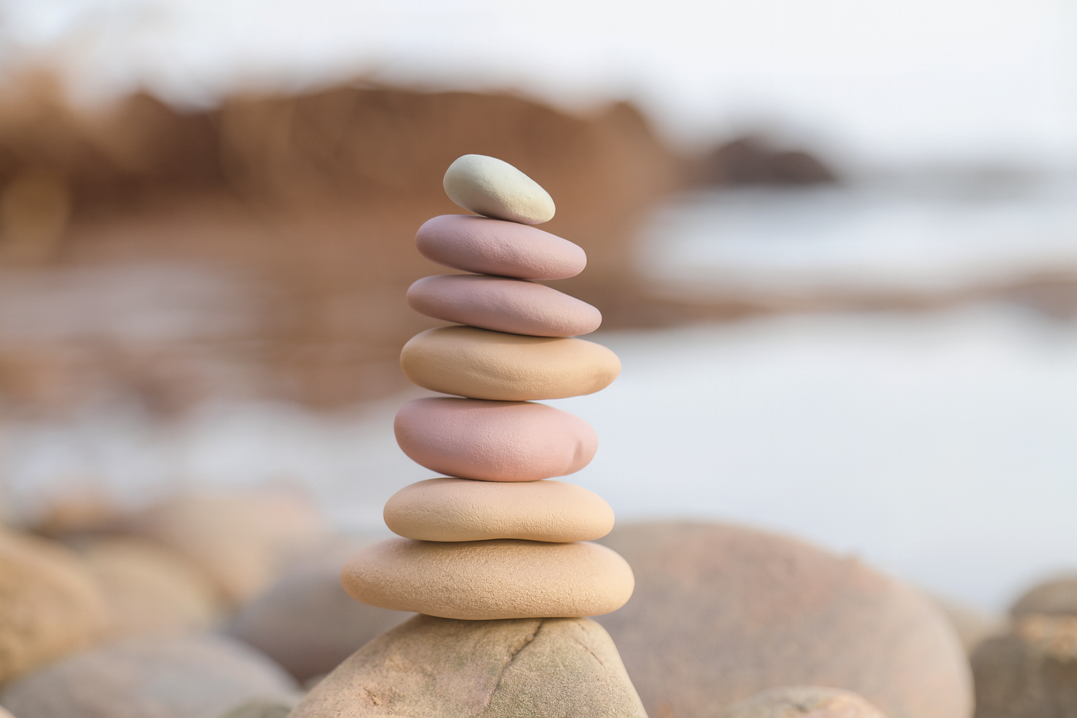 Stacked pastel-colored pebbles on a larger stone, with a blurred rocky shoreline and water in the background.
