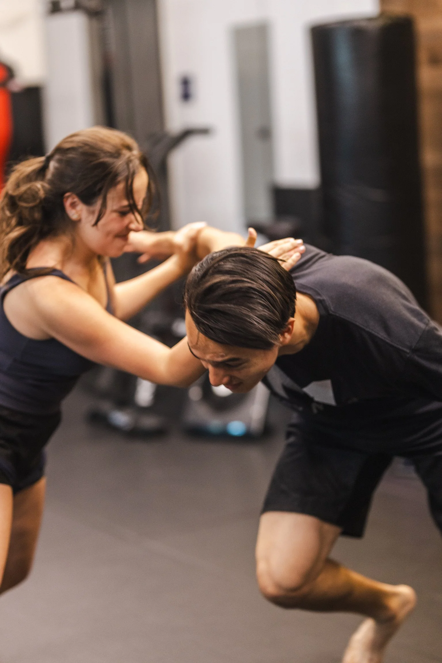 A woman practicing kickboxing with a male instructor in a gym, with her leg extended towards his midsection as he blocks it with his glove, both wearing athletic clothes and fighting gloves.