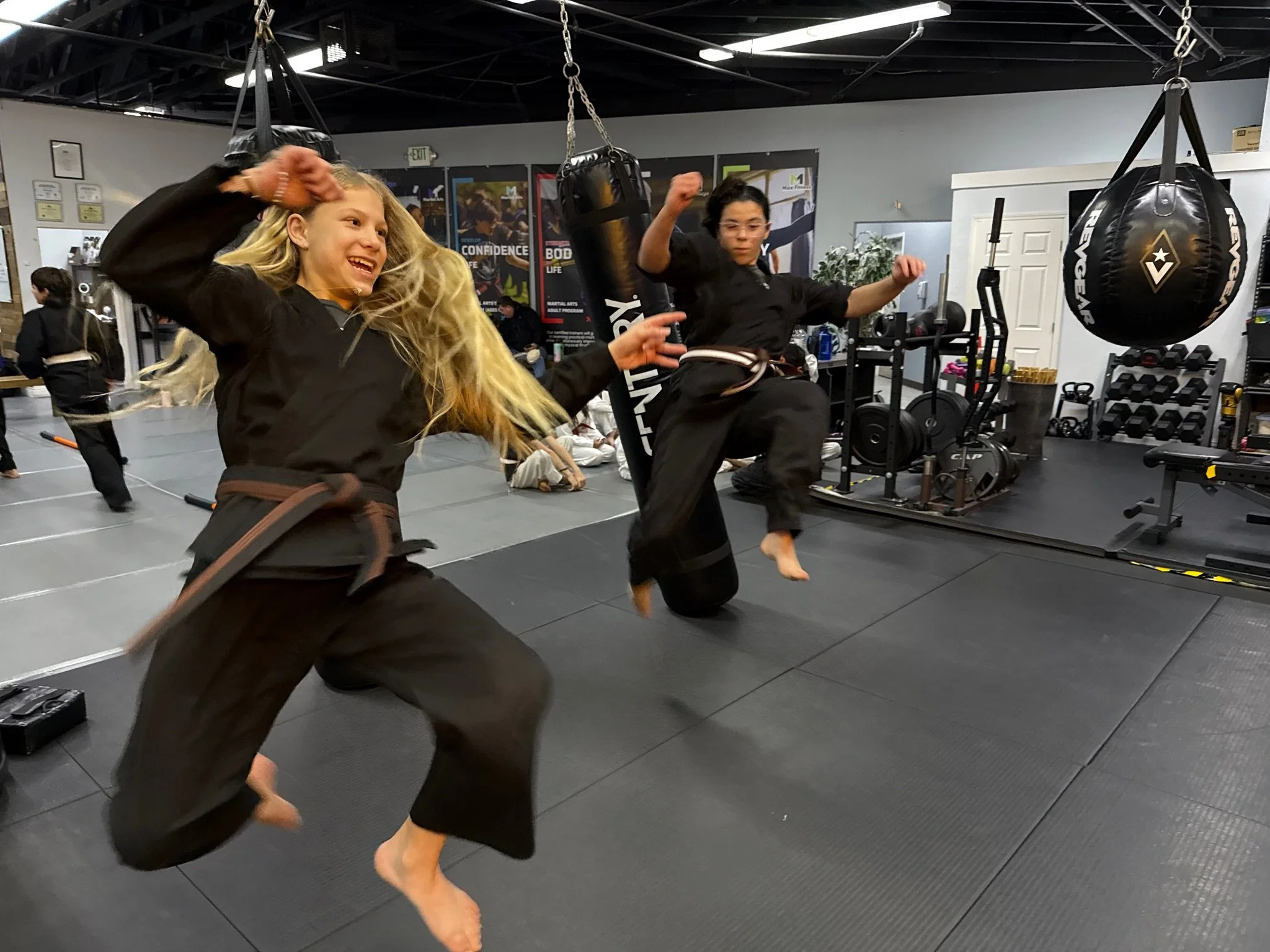 Two young girls practicing martial arts in a gym, with one girl wearing boxing gloves and the other with a training vest and holding a target pad, standing on a padded floor.