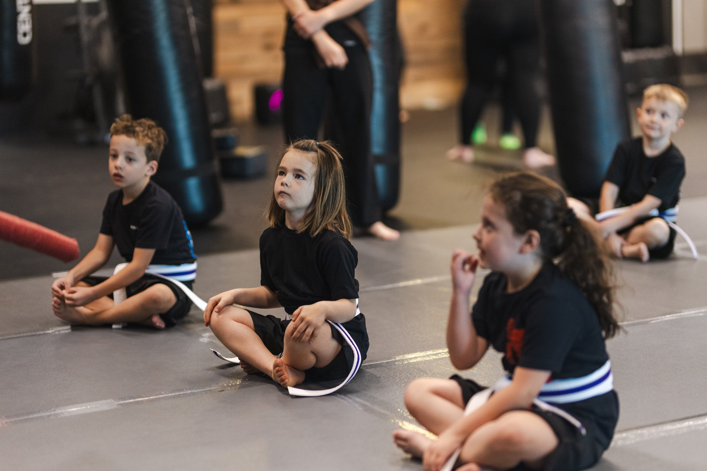 Children in martial arts uniforms standing and sitting on mats during a class at a martial arts gym.
