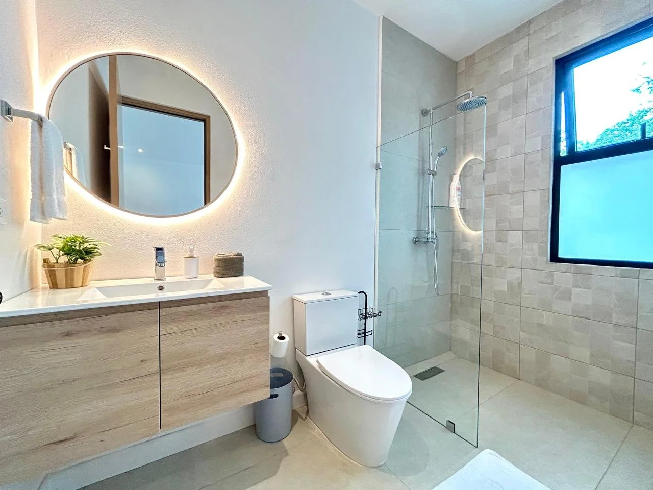 Un baño moderno con lavabo de madera clara, espejo redondo con luz de encendido, taza de baño blanca, ducha con mampara de vidrio y ventana grande con vista a árboles verdes.