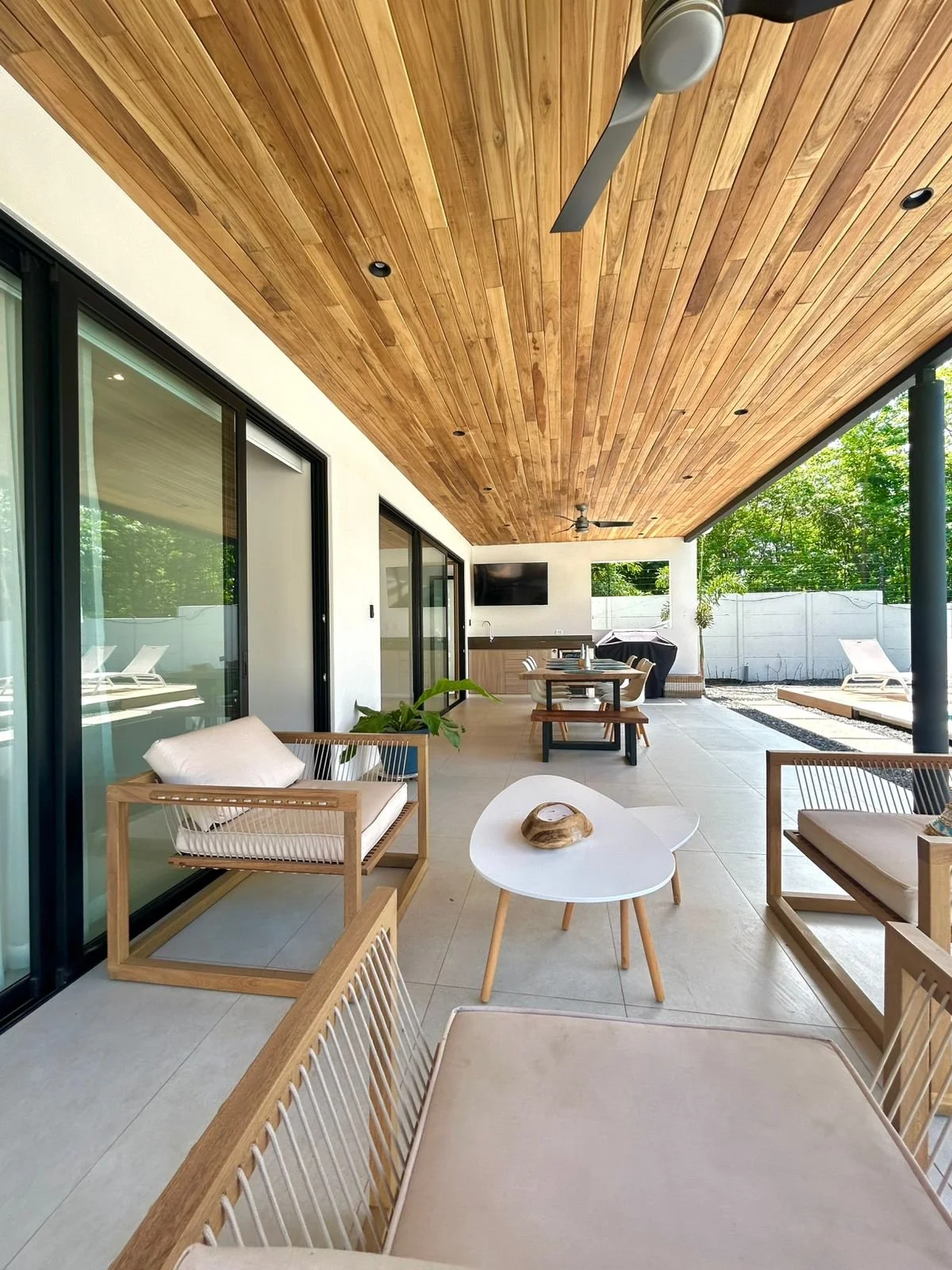 Terraza moderna con techo de madera, sillones de madera con cojines claros, mesa central blanca con adorno de madera, área de comedor con mesa y sillas de madera, televisión y área de descanso con tumbonas, paredes blancas y vista a un jardín con árb