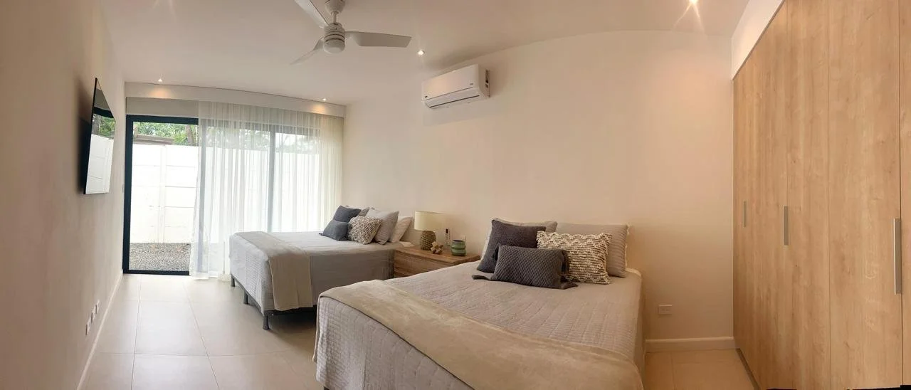 Dormitorio con dos camas, paredes blancas, armario de madera, ventanales con cortinas blancas, aire acondicionado en la pared.