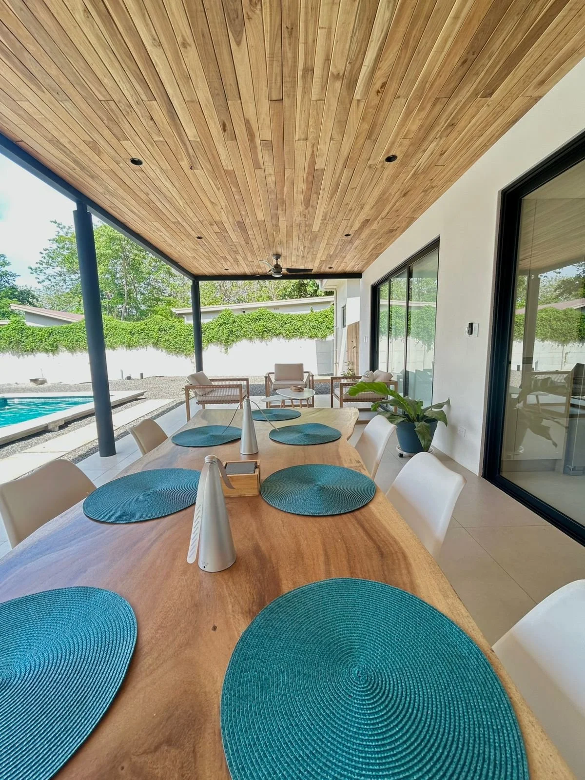 Vista del comedor en una terraza cubierta con mesa de madera y platos azules, con vista hacia el patios con plantas y piscina al aire libre.