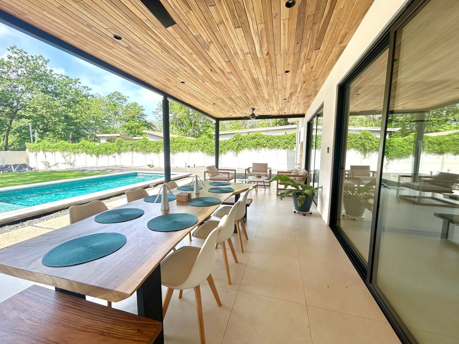 Comedor exterior con mesa larga, sillas blancas, y vista a piscina y jardín.