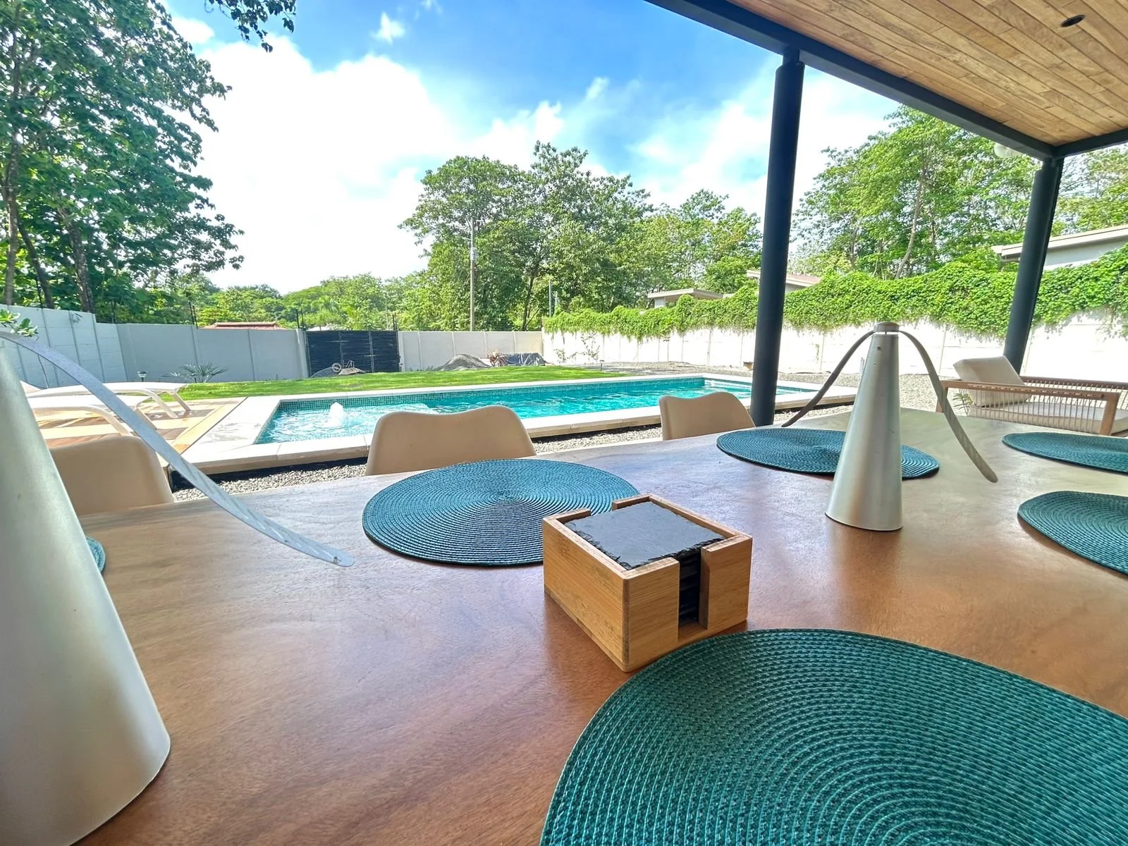 Mesa de madera con esquineros de tela azul, un objeto en forma de cono, y una caja de madera, vista hacia una piscina en un área con sombra y árboles verdes.