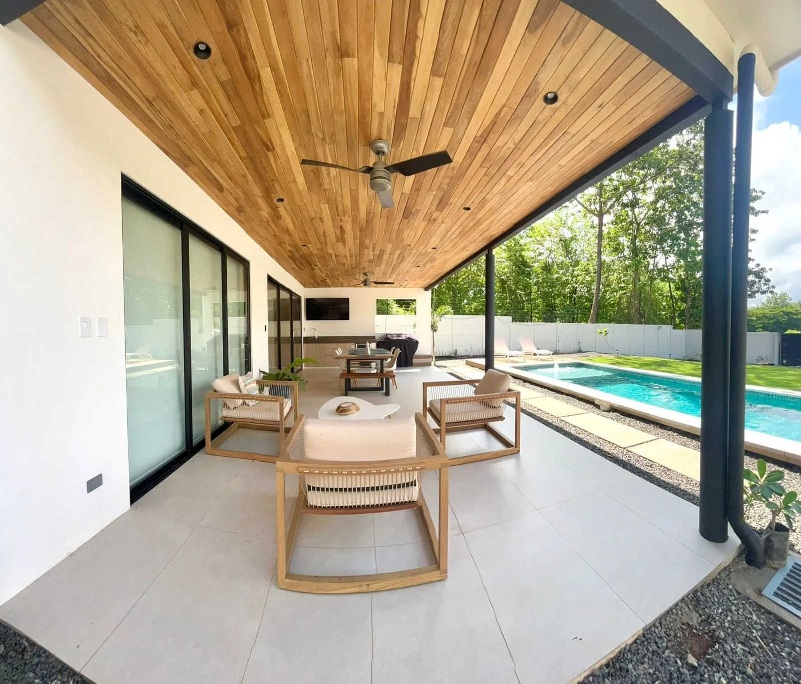 Ático con sillones de madera y cojines, área de comedor con mesa y sillas, piscina al aire libre, paredes blancas, techo de madera, ventiladores de techo, vista a un jardín con árboles.