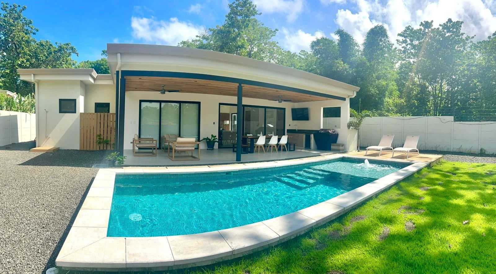 Casa moderna con piscina y área de descanso exterior, rodeada de árboles y césped.
