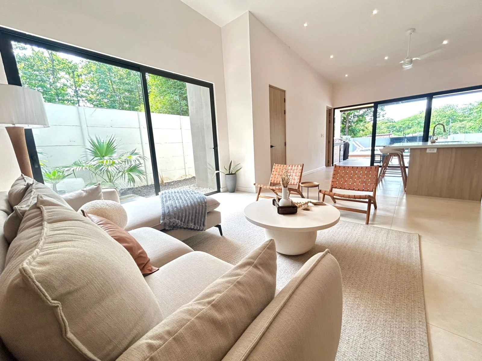 Sala moderna con sofá beige, mesa de centro blanca, sillas de mimbre, ventanas grandes con vista a árboles, plantas y zona exterior, iluminación moderna en techo.
