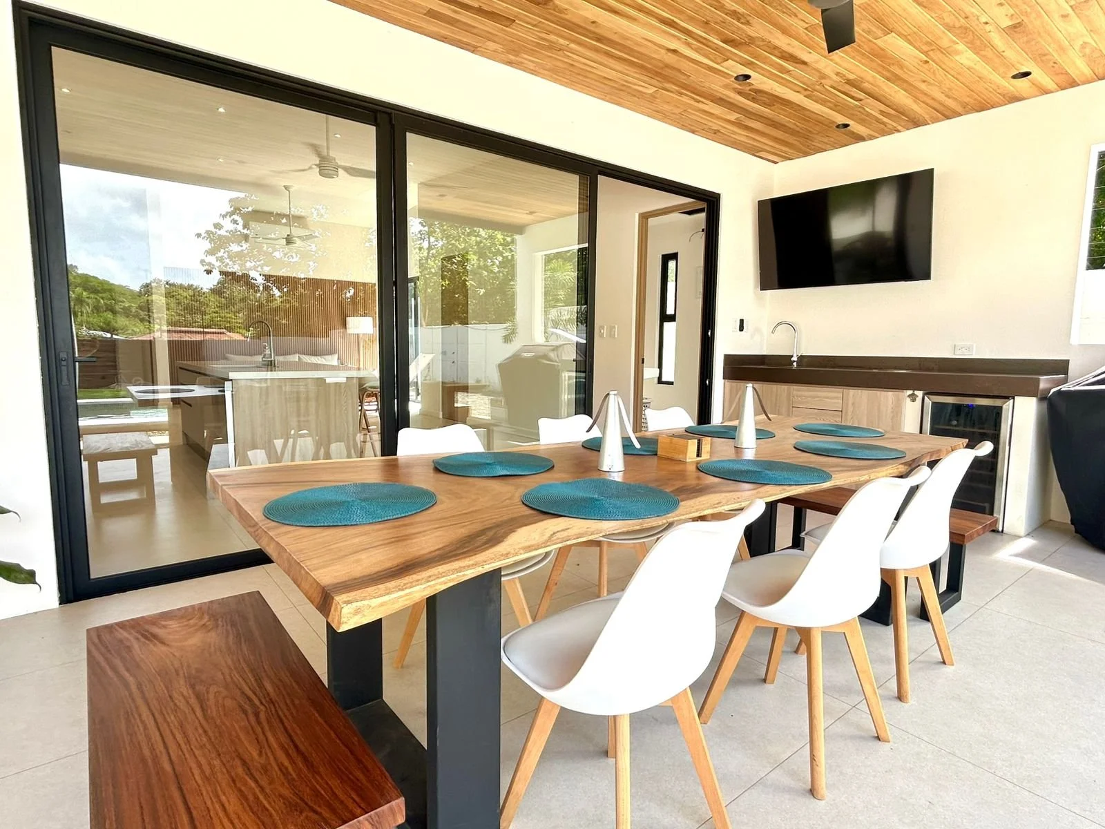 Comedor con mesa de madera, sillas blancas, decoraciones en tonos azul y vista a un espacio exterior mediante cortinas de vidrio.