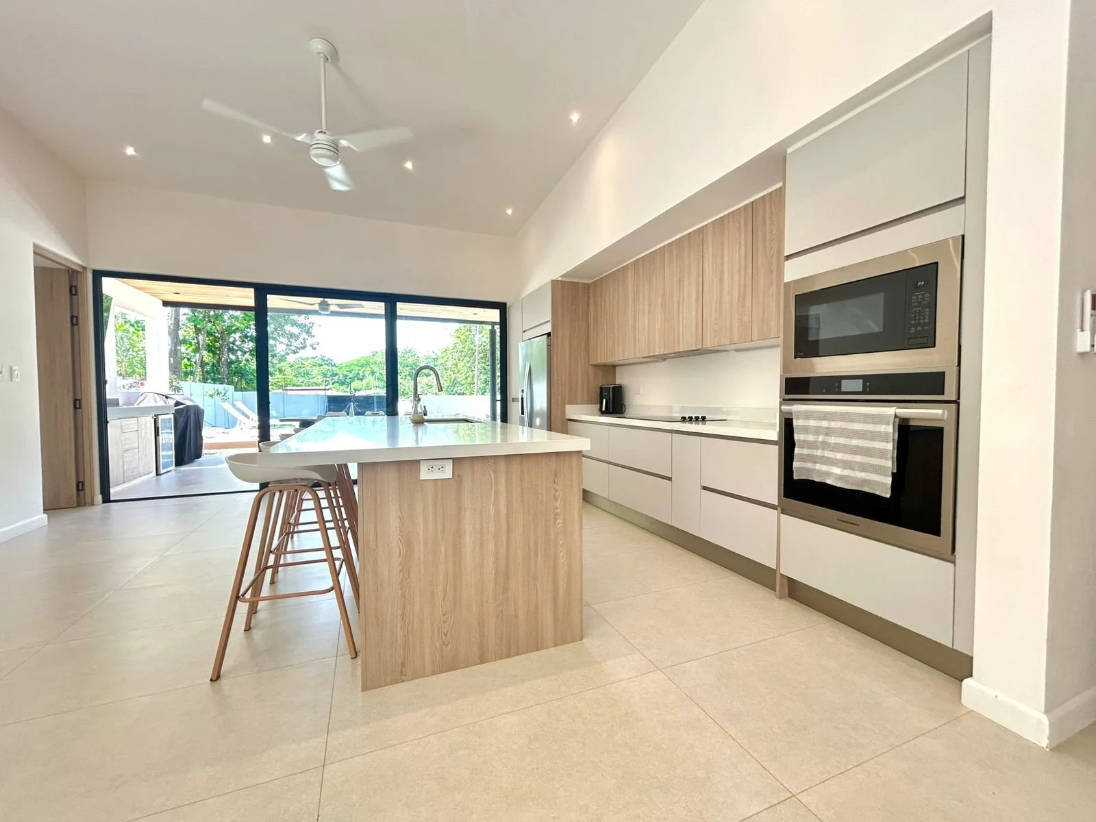 Cocina moderna con isla de encimera blanca, gabinetes de madera clara y electrodomésticos de acero inoxidable, con vista a un área exterior con árboles y espacio para taburetes en la isla.