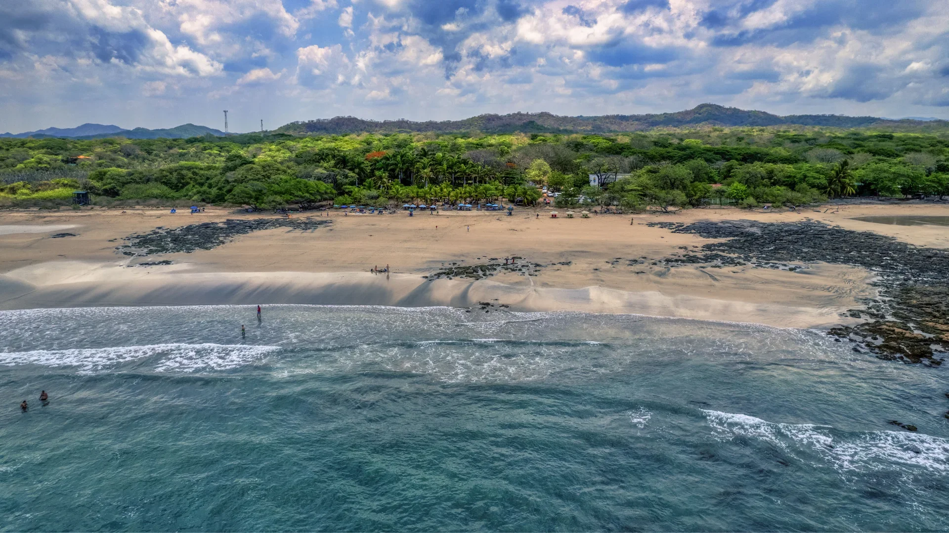 Playa con arena, rocas y personas en el mar, rodeada de vegetación y montañas con cielo nublado.