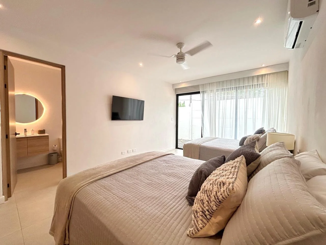 Habitación moderna con cama doble, televisor, aire acondicionado, y puerta hacia un patio con cortinas blancas.