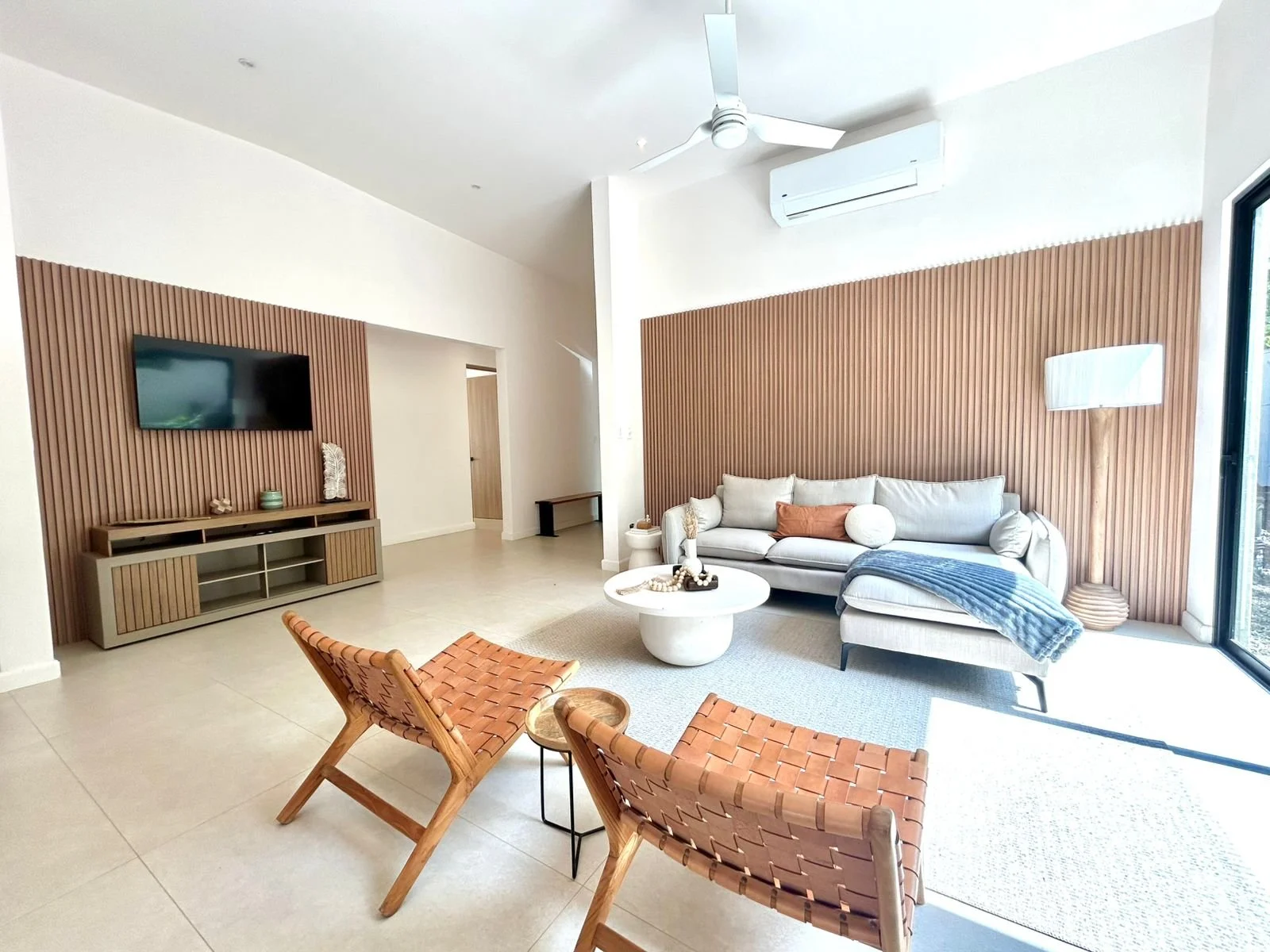 Sala de estar moderna con pared de madera, sofá gris con cojines, una lámpara de pie, sillas de madera con asiento de cuero entre una mesa redonda y una ventana grande con vista al exterior
