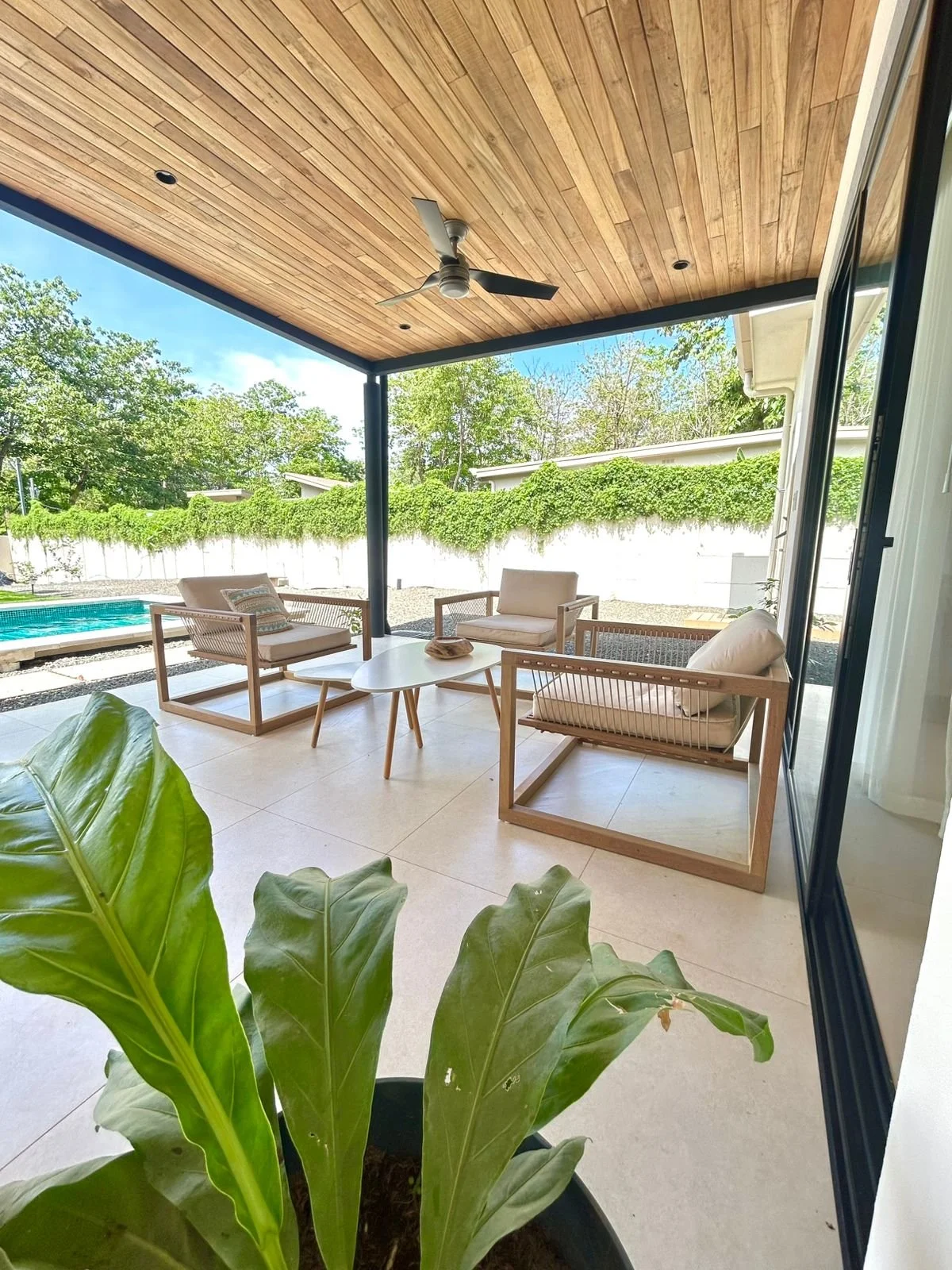 Terraza cubierta con techo de madera, ventilador de techo, mobiliario de sala con sillones y mesa, cercado con árboles y jardín en el fondo, planta en primer plano.
