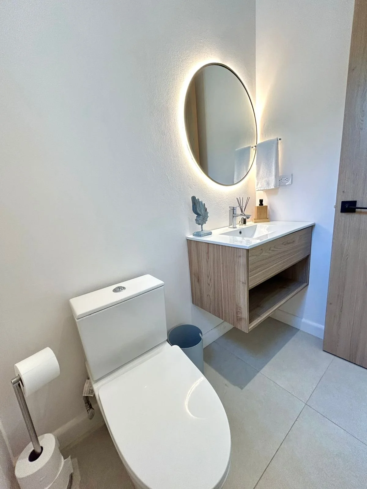 Un baño moderno con sanitario blanco, lavabo de madera con espejo circular y luz de fondo, accesorios de tocador y cortina de toallas en una pared blanca.