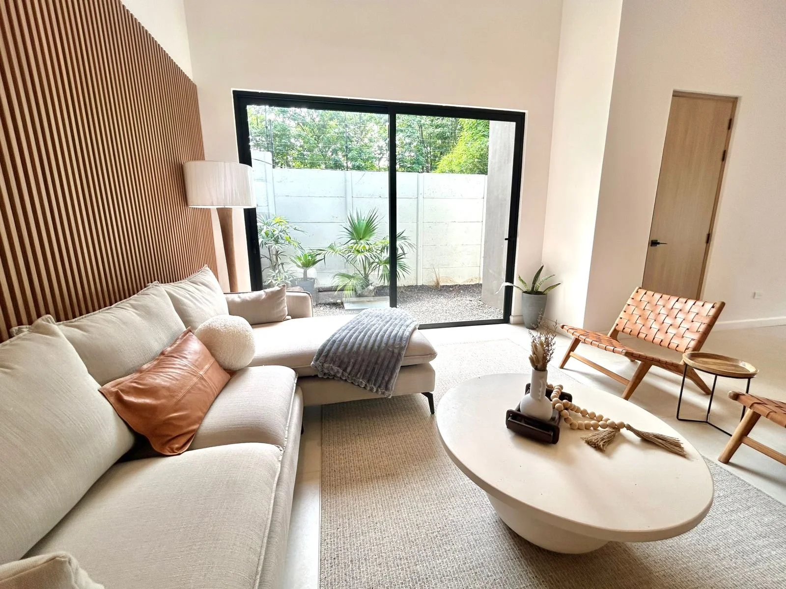 Sala moderna con sofá beige, mesa de centro ovalada, sillas de mimbre, y planta en maceta junto a ventana de vidrio grande que da a un patio con plantas.