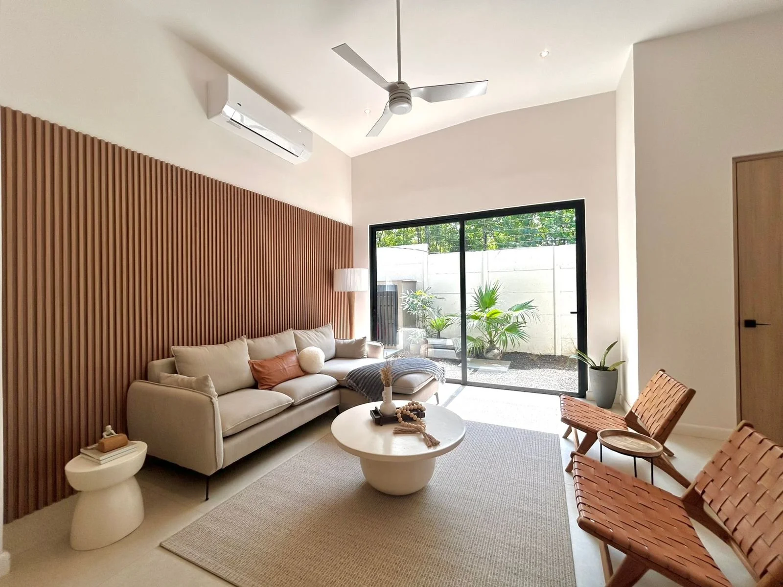 Sala moderna con sofá beige, mesita redonda y sillones de madera, ventanales con vista a jardín con plantas verdes, paredes blancas, techo con ventilador y aire acondicionado.