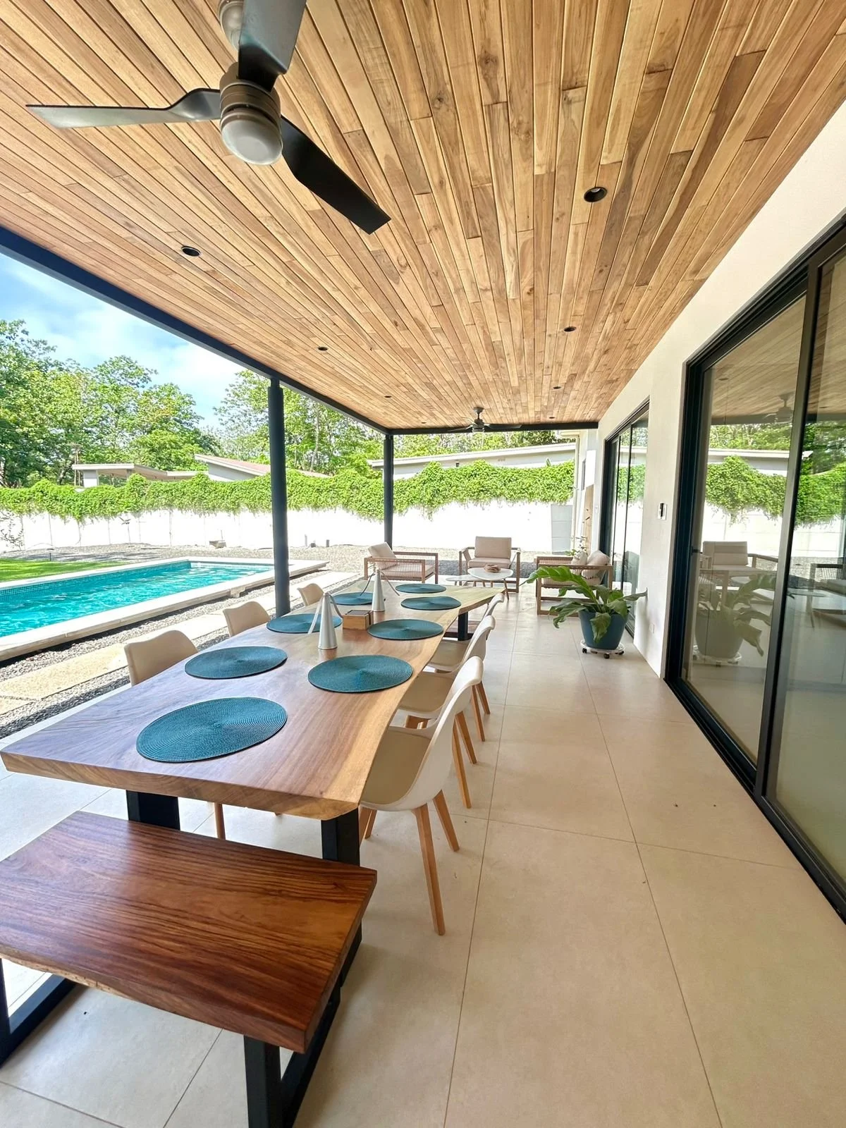 Espacio de comedor al aire libre con mesa de madera, sillas blancas, y decoración moderna, con vista a la piscina y áreas verdes.