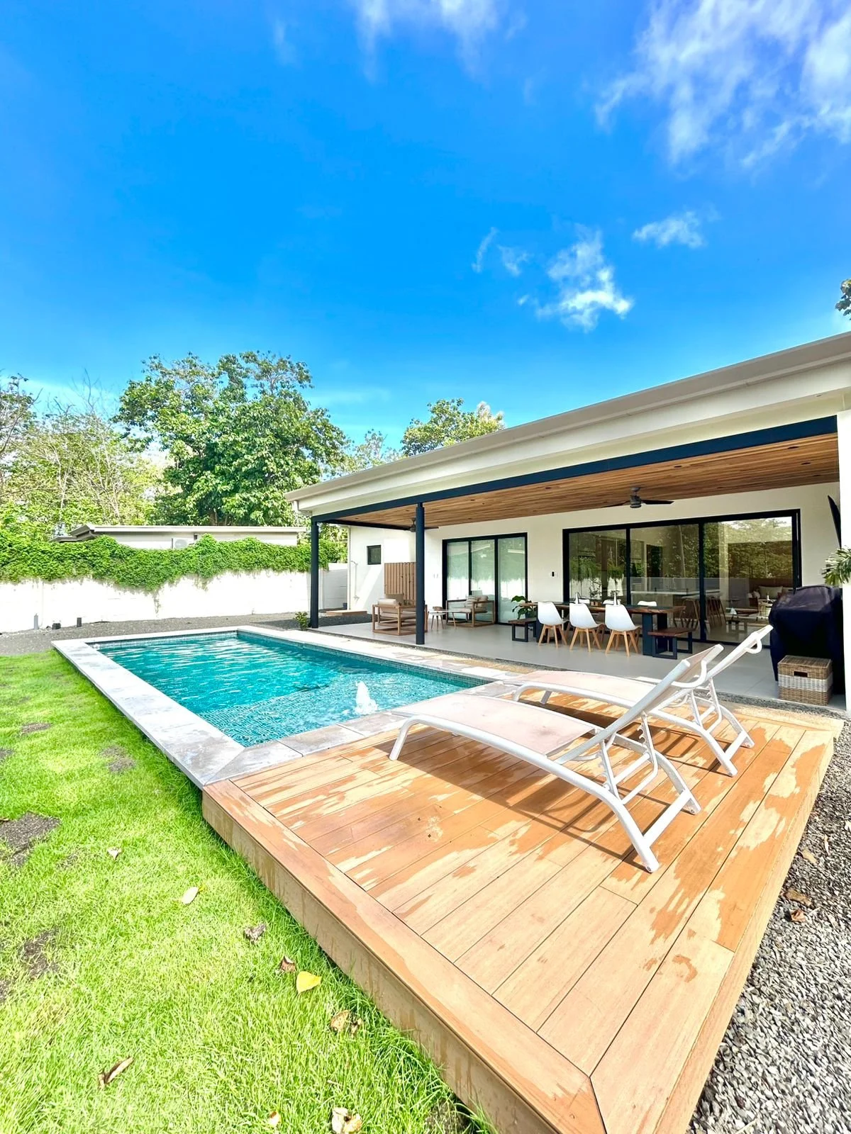 Piscina en el patio trasero de una casa moderna con muebles de exterior, césped verde, un área de descanso y una casa con ventanas de vidrio