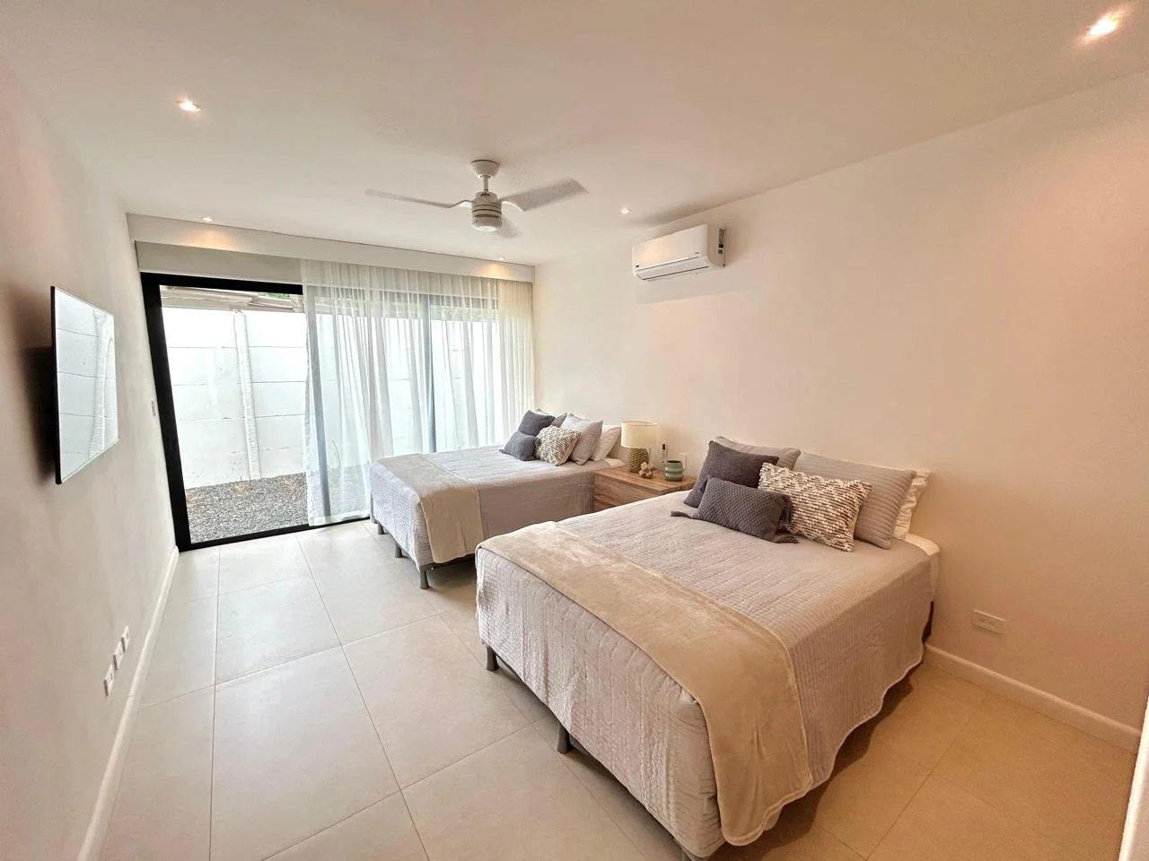 Dormitorio con dos camas, sábanas claras y múltiples almohadas, vista a la puerta corrediza con cortinas blancas, aire acondicionado en la pared y televisor en la pared.