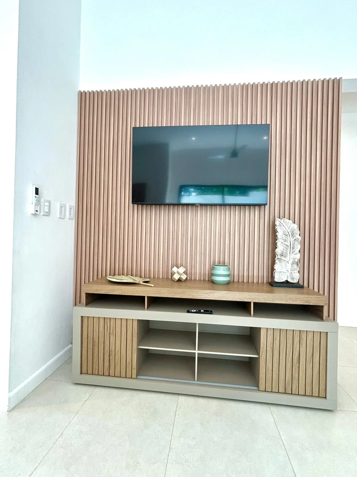 Mueble de madera con espacio para almacenamiento en la parte inferior, sobre un suelo claro, con una pared de paneles de madera en la parte trasera y un televisor montado en ella, además de decoraciones como una hoja seca, una figura decorativa en fo