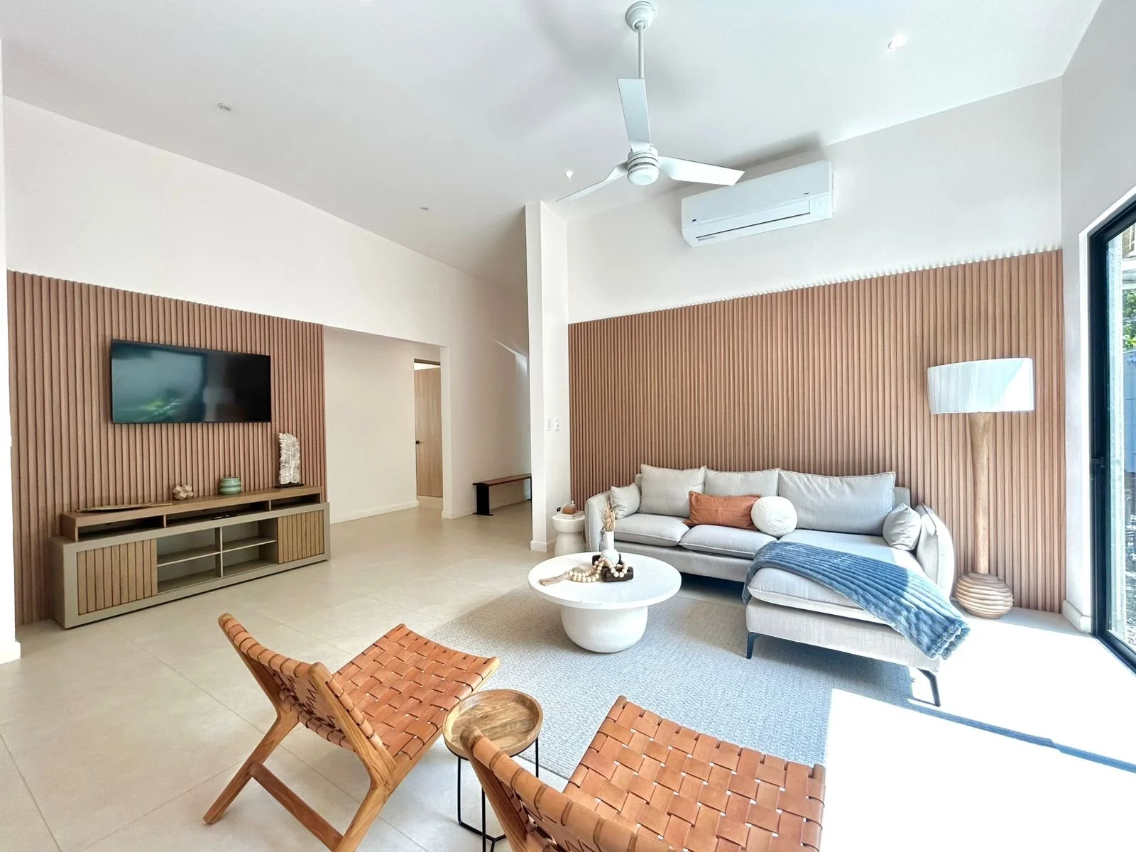 Sala de estar moderna con pared de madera, sofá gris con cojines, mesa de centro blanca, lámpara de piso, televisión en la pared y ventilador en el techo, con entrada a un espacio exterior con ventana grande.