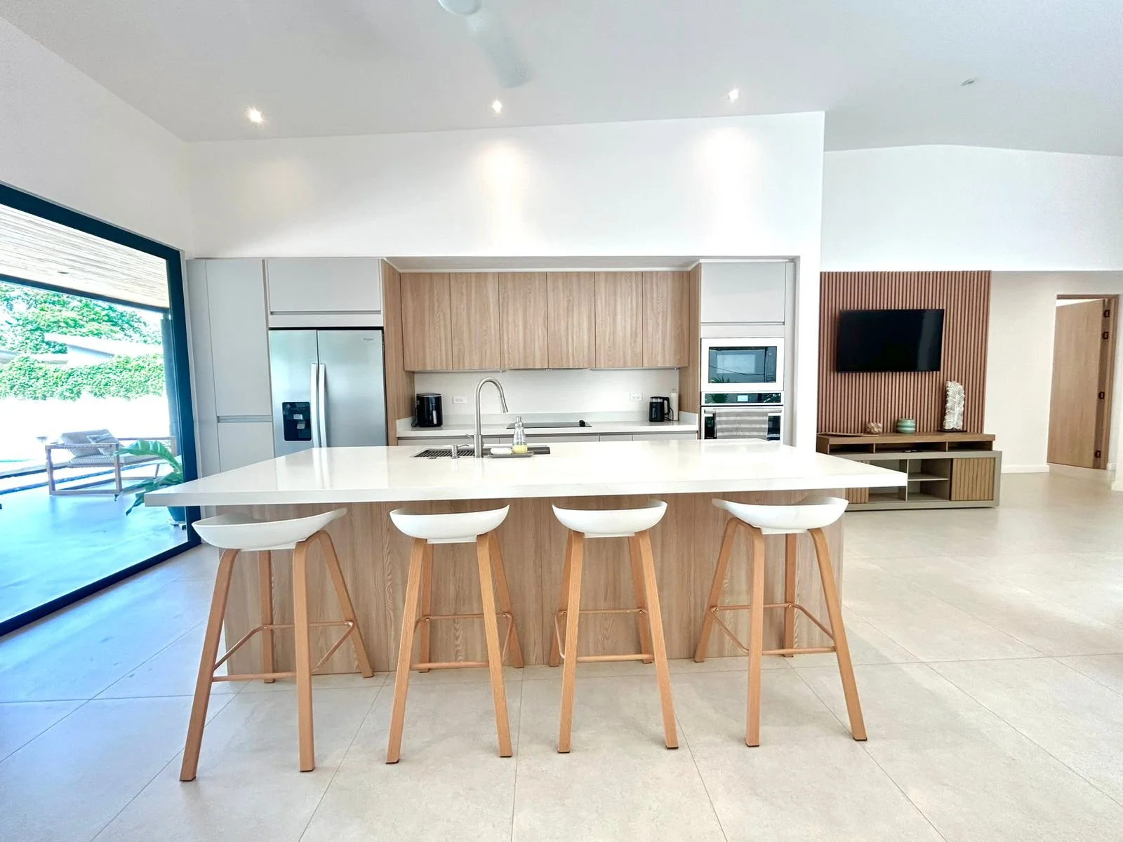 Cocina moderna con isla central de madera y bancos blancos con patas de madera natural, electrodomésticos integrados, vista a un amplio espacio y televisión en la pared
