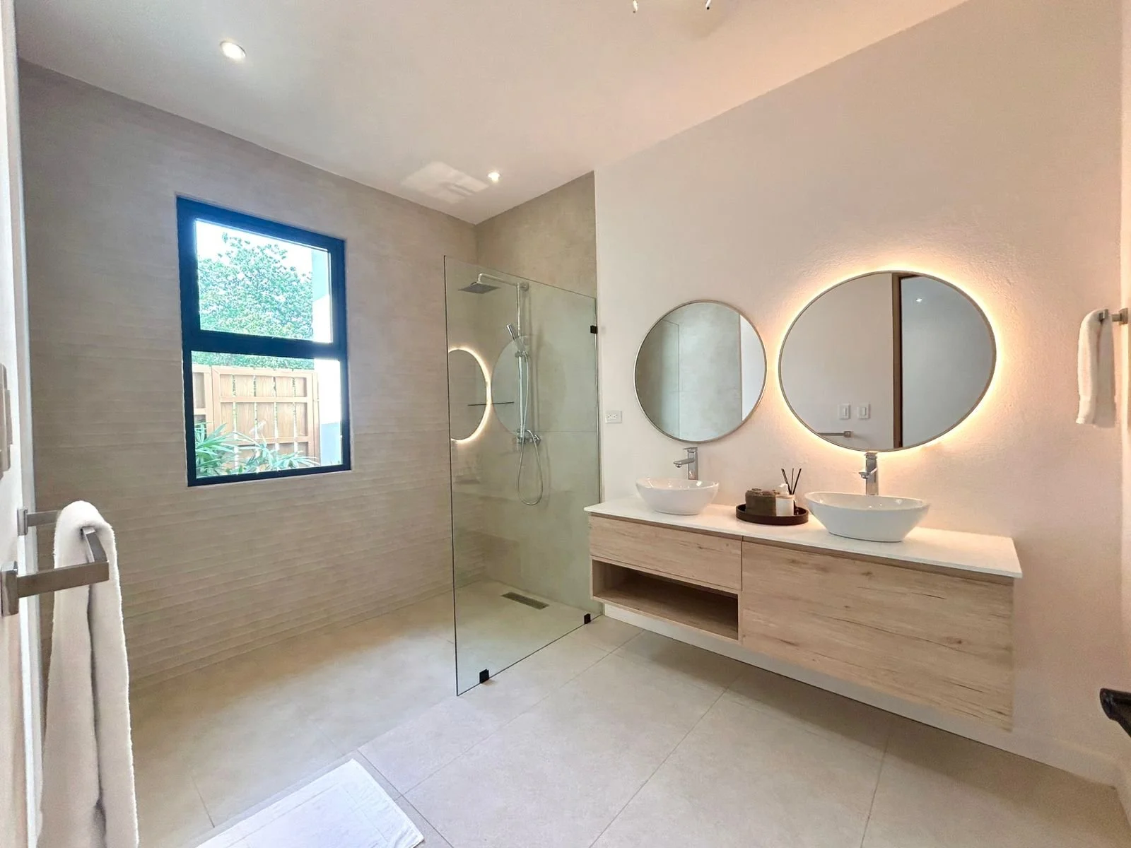 Baño moderno con doble lavabo con espejos de iluminación led, área de ducha con puertas de vidrio, ventana grande y decoraciones sencillas.