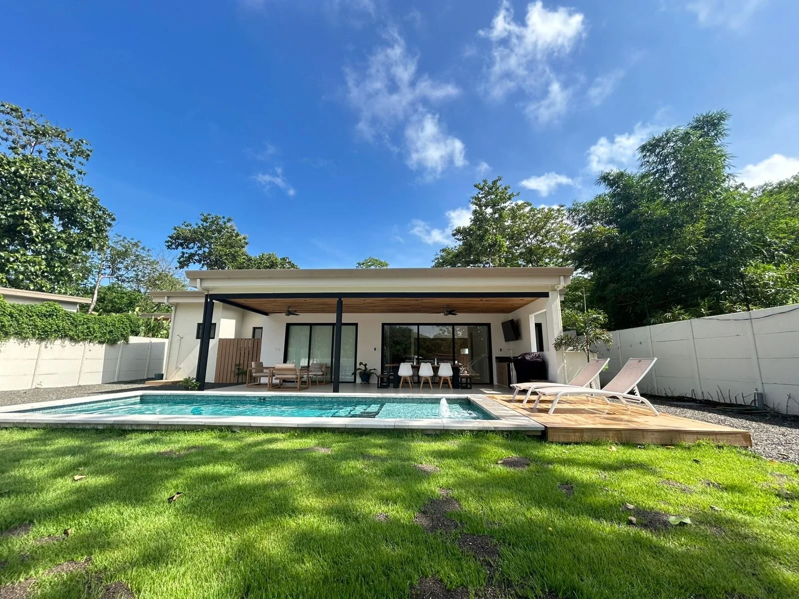 Casa moderna con piscina y zona de descanso en un jardín con césped bajo un cielo azul con nubes.