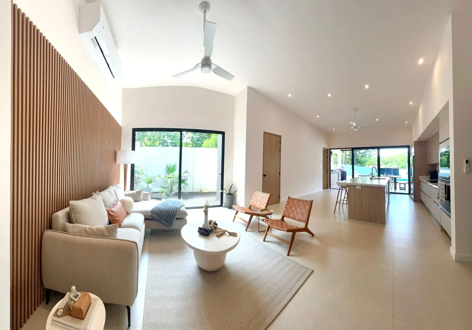 Sala moderna con sofá beige, sillones de madera, mesa de centro, lámpara de pie, pared de madera, puerta de vidrio que da al patio, cocina con isla y vista al jardín, pisos claros, iluminación empotrada y aire acondicionado.