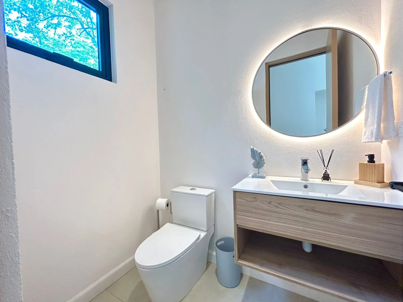 Baño moderno con inodoro blanco, lavabo de madera con espejo redondo con luz, ventana azul y accesorios decorativos en el lavabo.