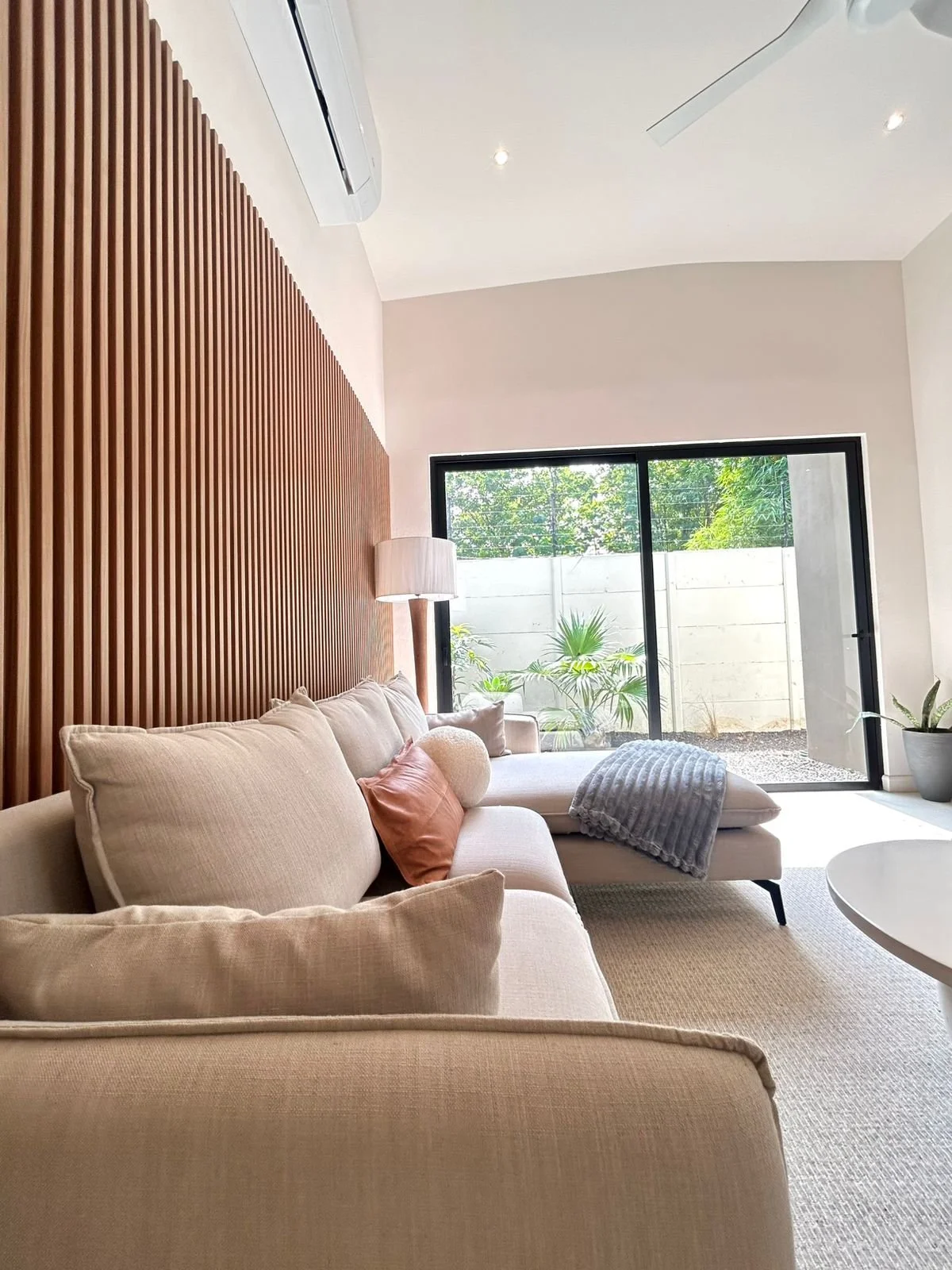 Sala moderna con sofá beige, cojines en tonos tierra, una lámpara de pie, ventana grande con vista a un jardín con plantas