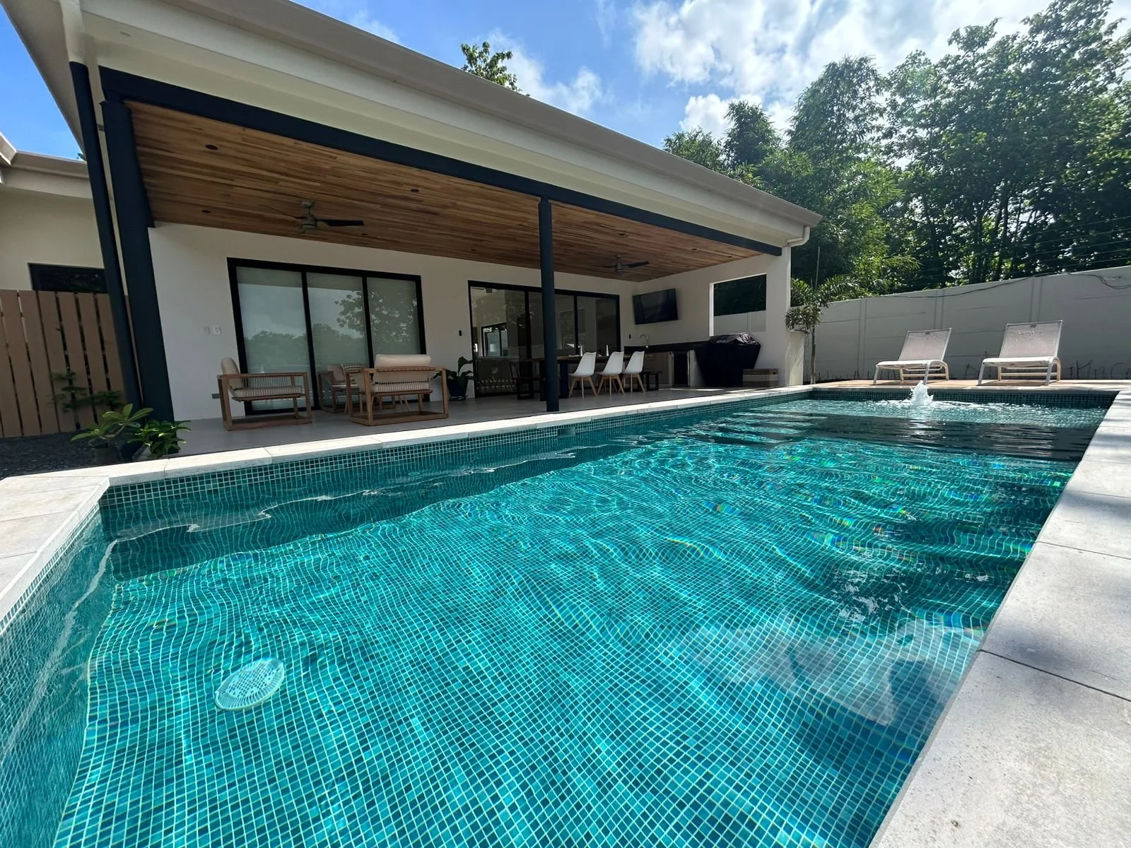 Piscina en un patio trasero con mobiliario y una casa moderna de fondo, rodeada de árboles y cielo azul.