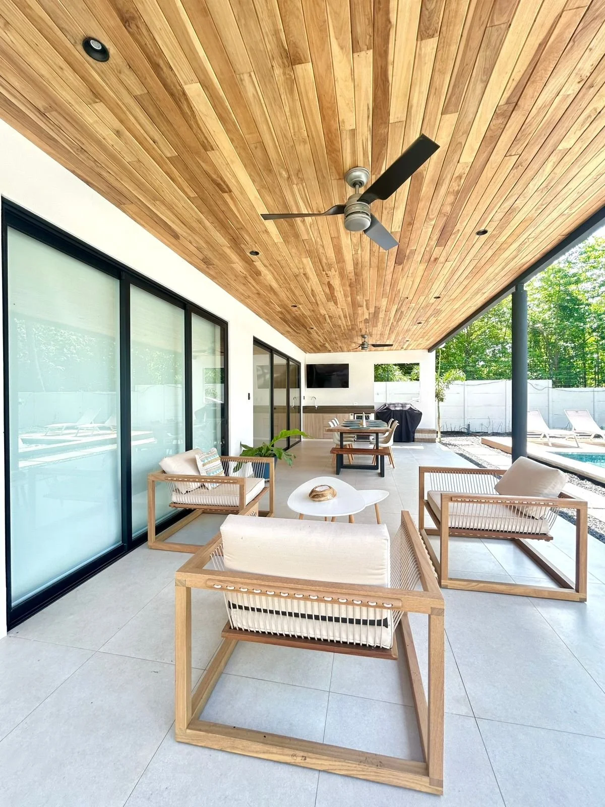 Terraza al aire libre con muebles de madera y cojines, techo de madera con ventiladores, paredes de vidrio con cortinas, piscina y área de descanso con vistas a la naturaleza.