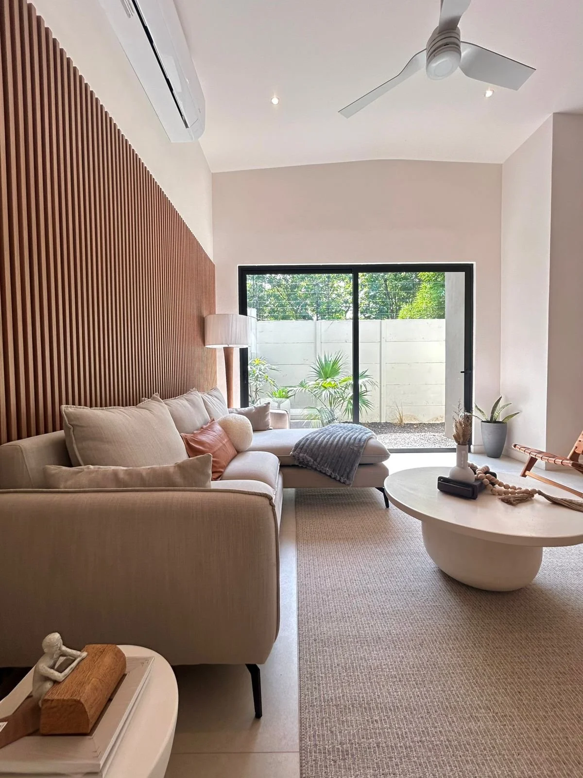 Sala de estar moderna con sofá beige, mesa de centro redonda y puertas corredizas con vista al patio con plantas