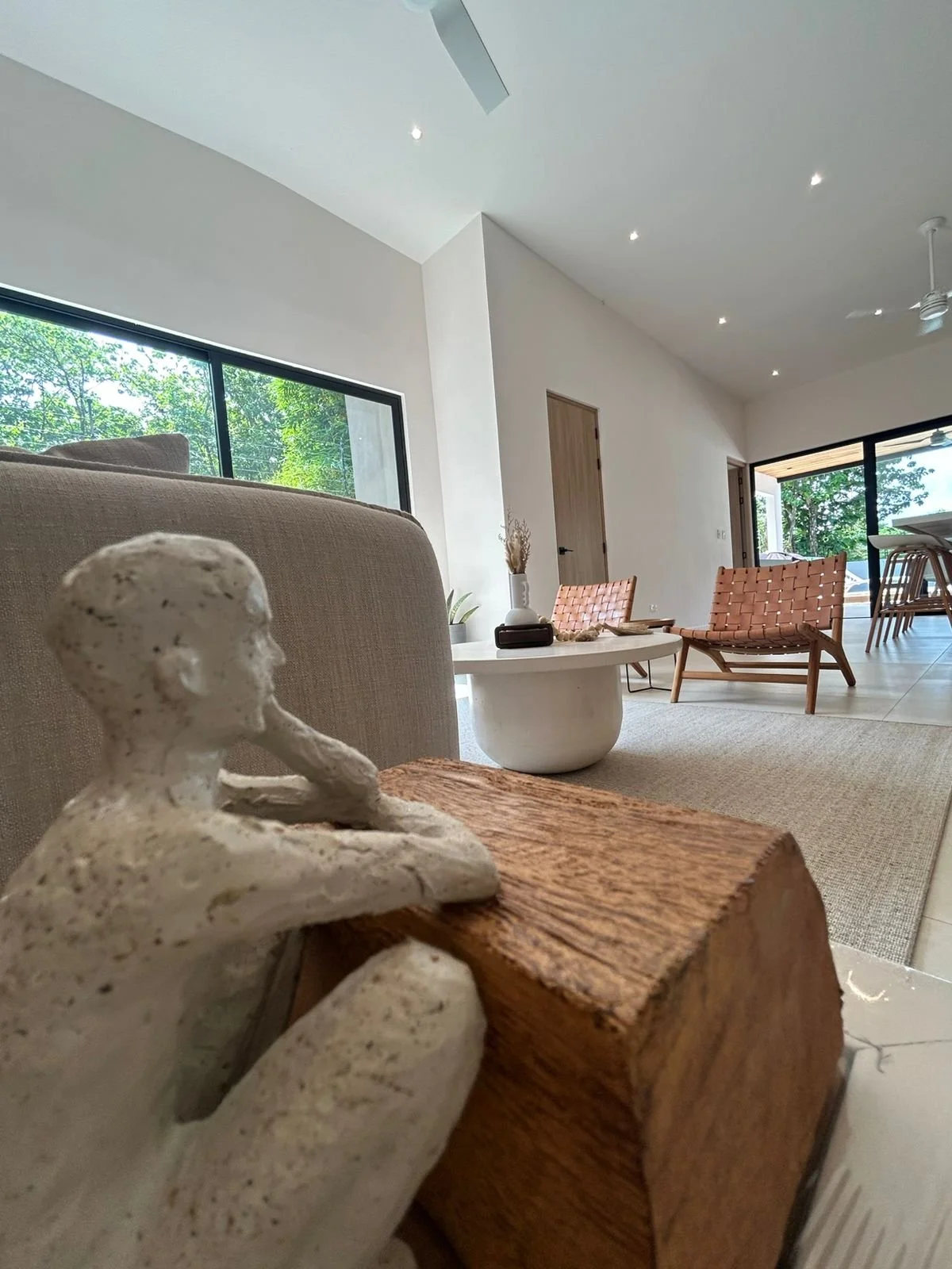 Vista del interior de una sala con muebles de madera y decoraciones modernas, incluyendo una escultura de un hombre, una mesa blanca y sillones de piel de color marrón, con grandes ventanas que dejan entrar luz natural y muestran árboles verdes afuer