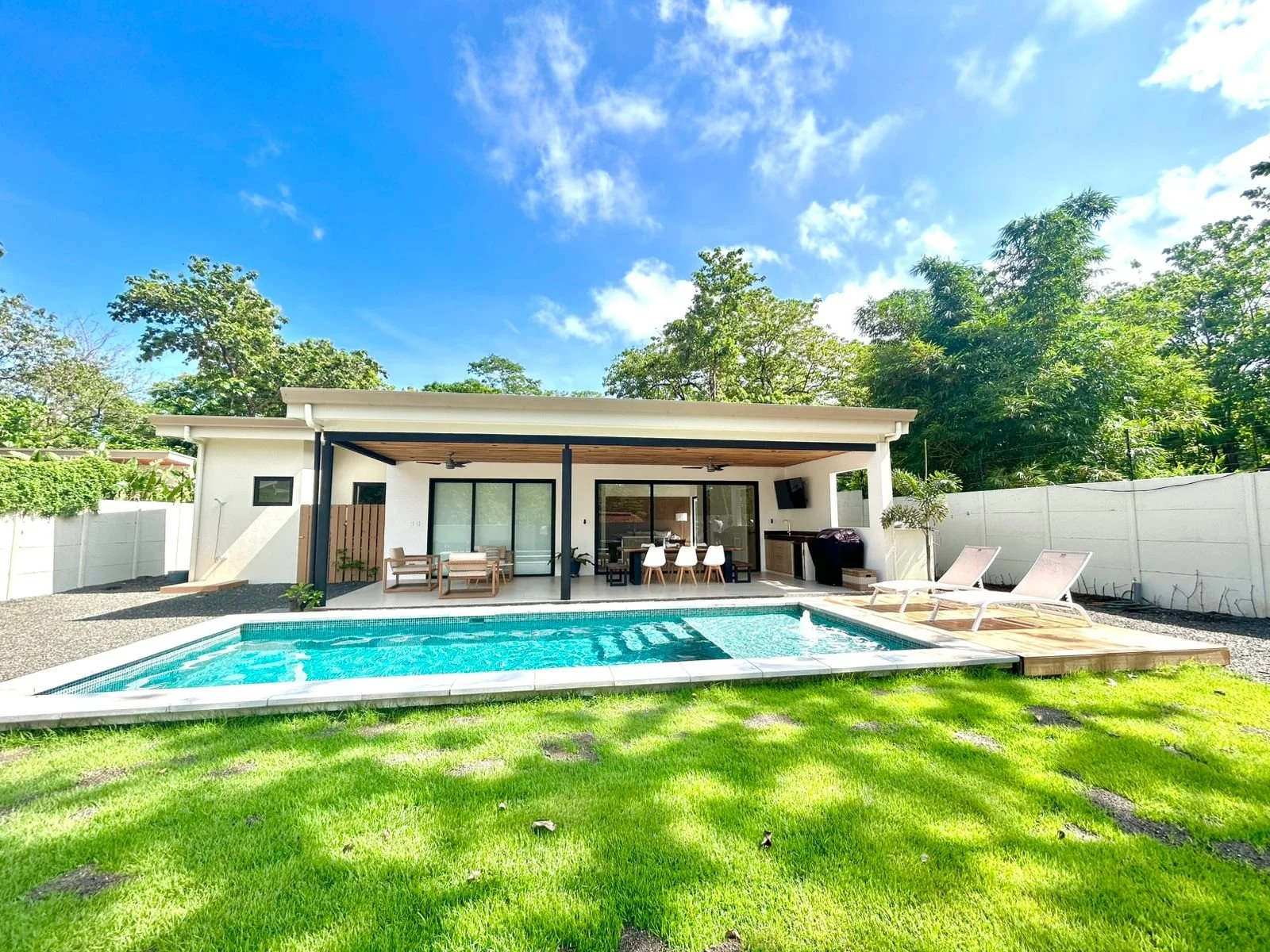 Casa moderna con piscina y área de descanso, rodeada de árboles y césped, en un día soleado con cielo azul.