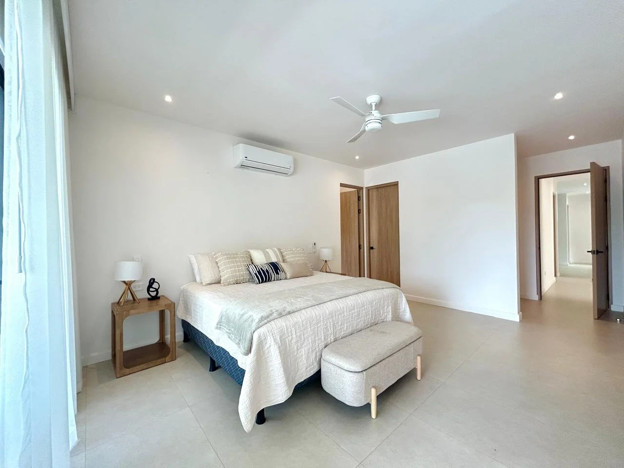 Habitación moderna con cama grande, ropa de cama blanca, mesitas de noche con lámparas, ventilador de techo, aire acondicionado, puertas de madera y suelos claros.
