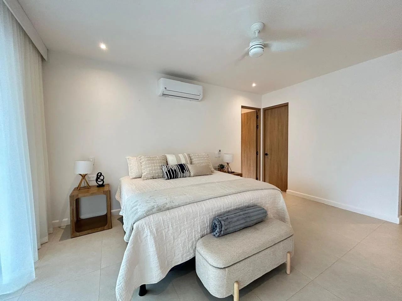 Habitación moderna con cama grande, colchas blancas, varios cojines, lámparas de noche, una ventana grande con cortinas, aire acondicionado y ventilador en el techo, pisos de cerámica y puertas de madera.