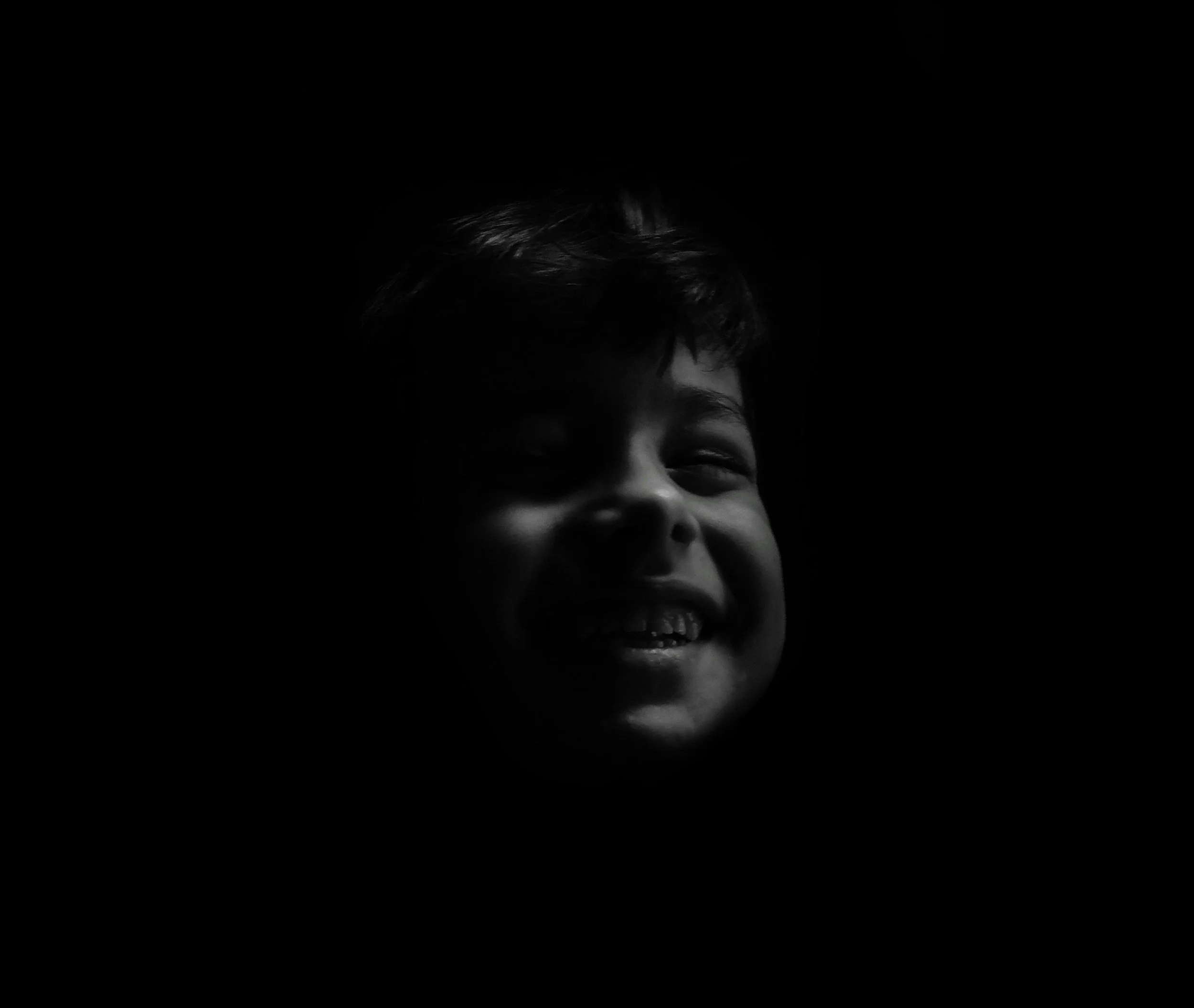 Black and white portrait of a smiling child with short hair, illuminated softly on the face against a dark background.